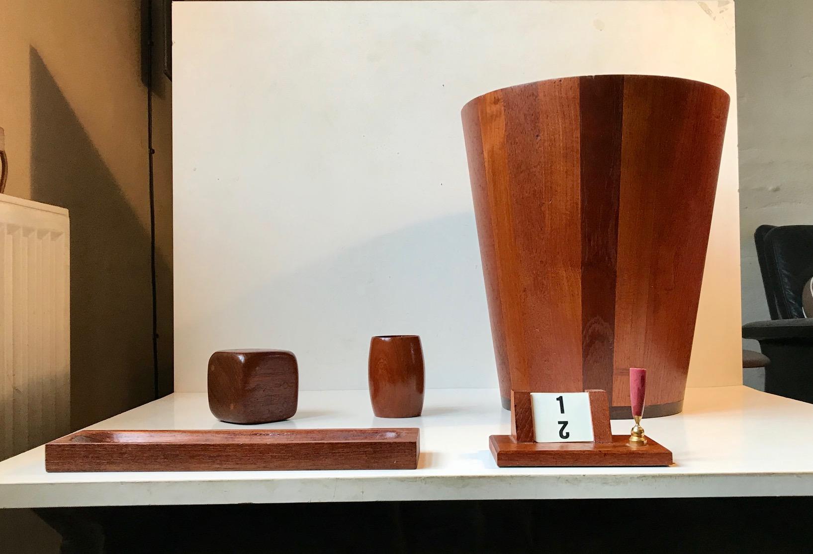 A set of desk/office accessories consisting of af solid stacked teak waste basket, a dice paperweight, pen tray, pen holder with calendar and a vase/container for clips etc. Manufactured from leftover teak from different Danish furniture makers