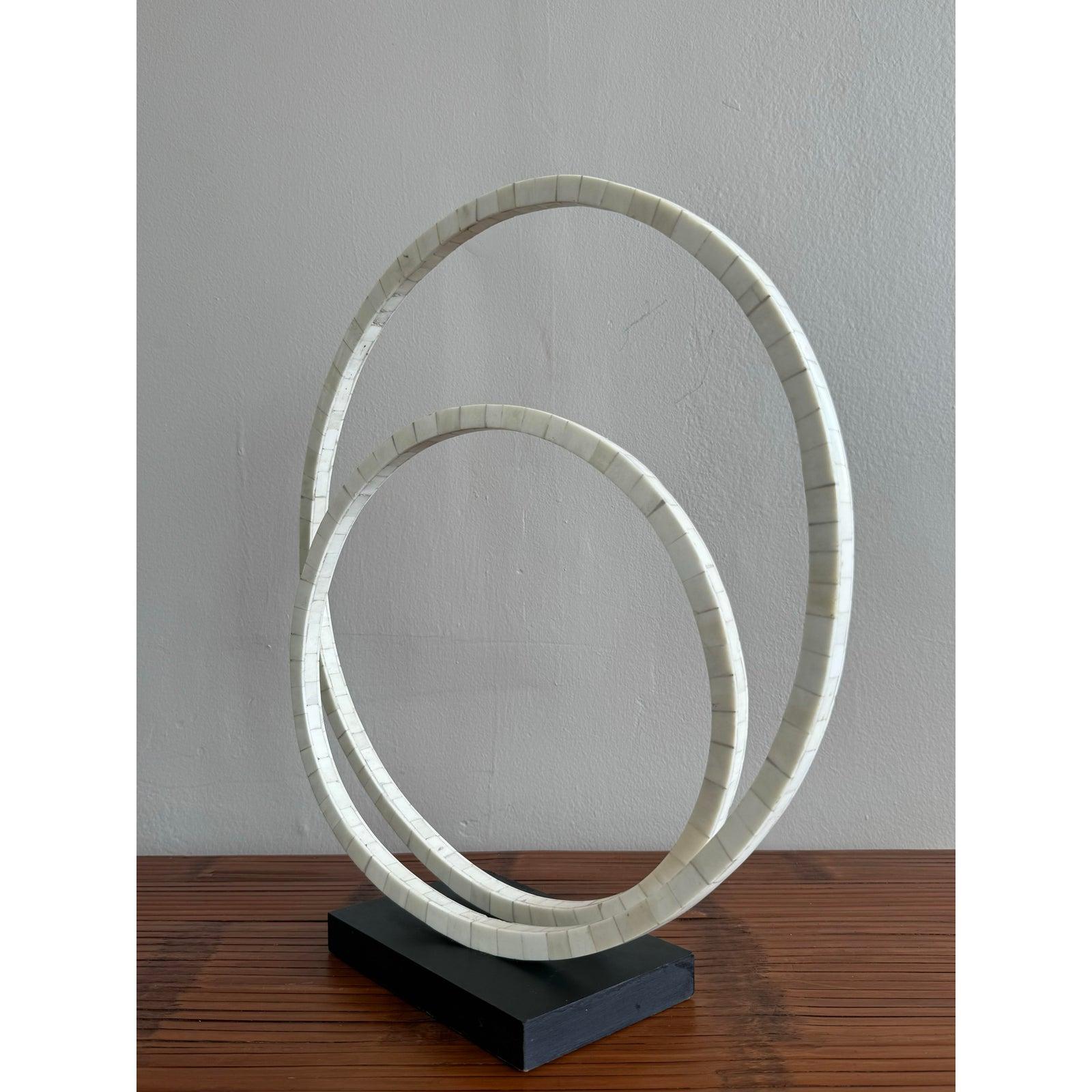 Questa scultura astratta presenta una coppia di forme ellittiche ad anello rivestite con una finitura liscia di colore avorio, poste su una solida base nera. Il gioco dei due anelli crea un ritmo tranquillo che suggerisce movimento, continuità o