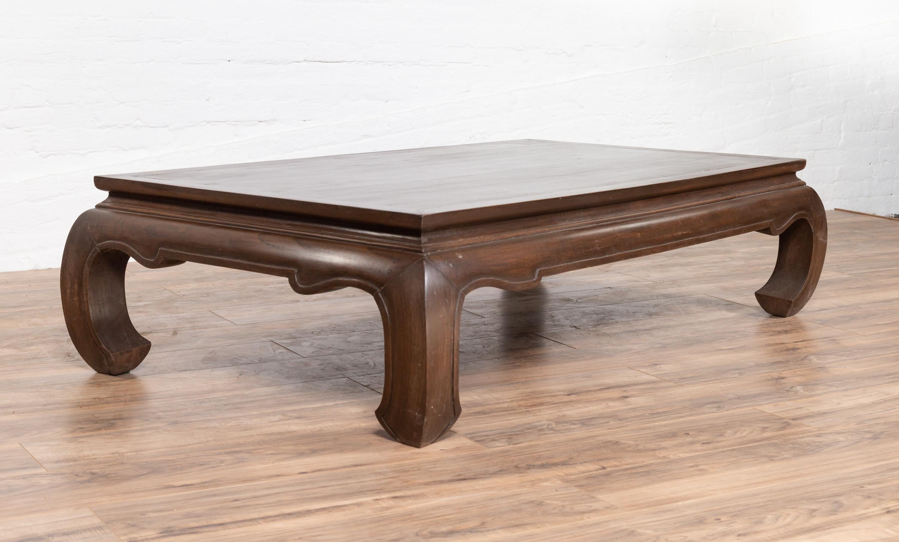 A vintage Thai opium bed style coffee table from the mid-20th century with dark patina and chow legs. Born in Thailand during the mid-century period, this elegant coffee table features a rectangular top sitting above a waisted apron with curving