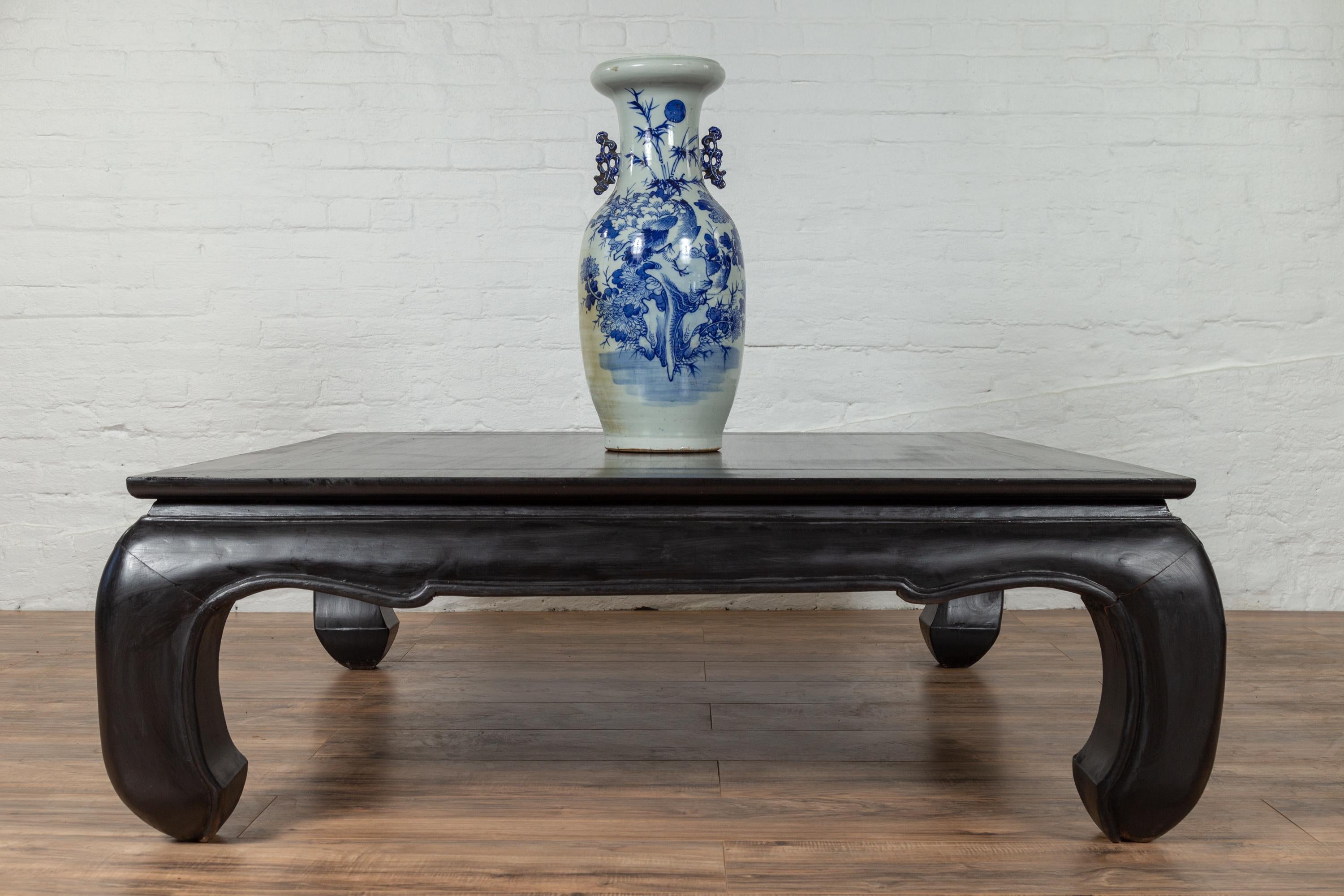 A vintage Thai teak coffee table from the mid-20th century, with large chow legs and black lacquer. Born in Thailand during the midcentury period, this teak coffee table features a square planked waisted top sitting above an ornamental curving