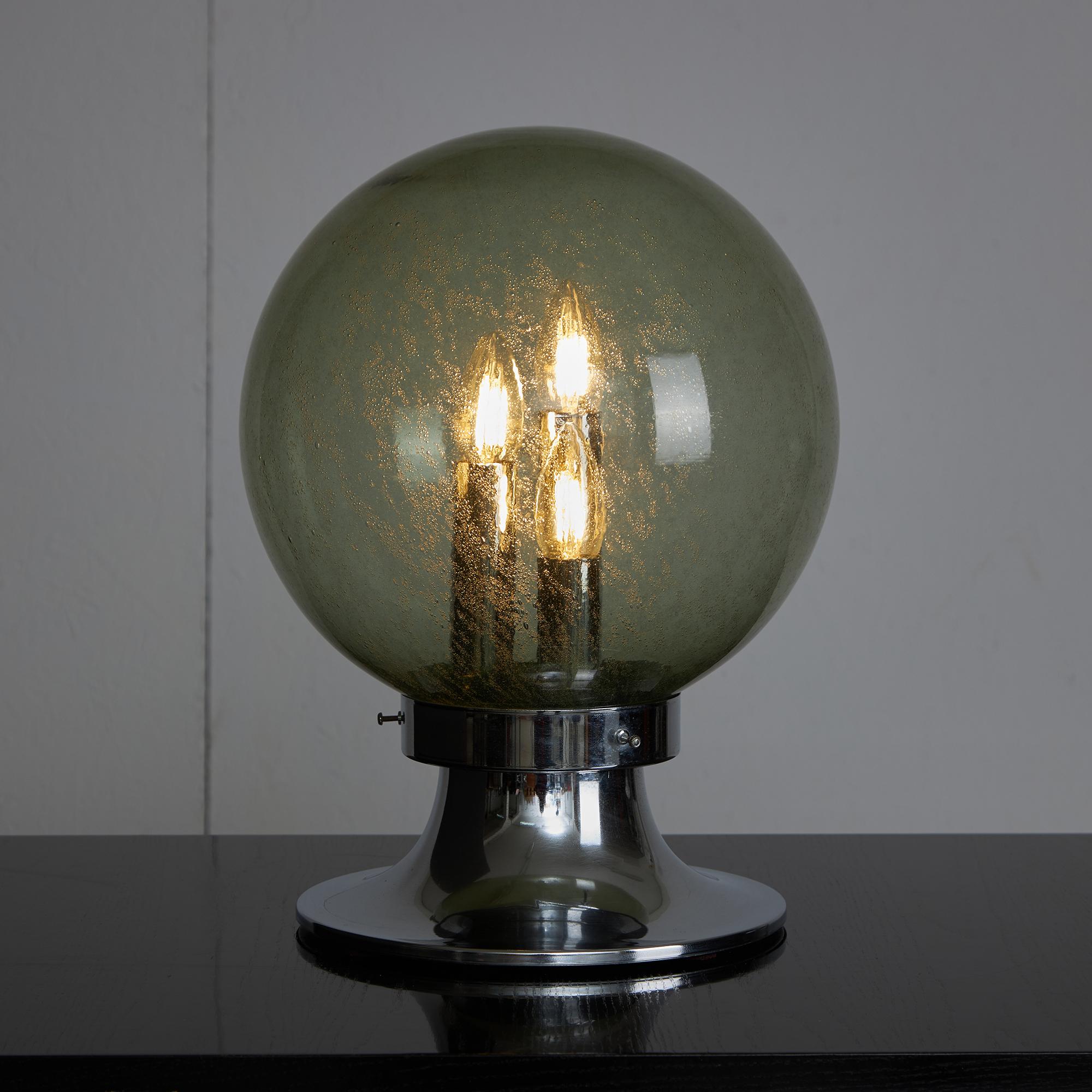 A one-of-a-kind table lamp featuring a glass globe shade adorned with delicate bubbles, adds an enchanting texture to its design. Inside, three bulbs are cleverly concealed, resembling emerging chrome towers that catch the light beautifully. The