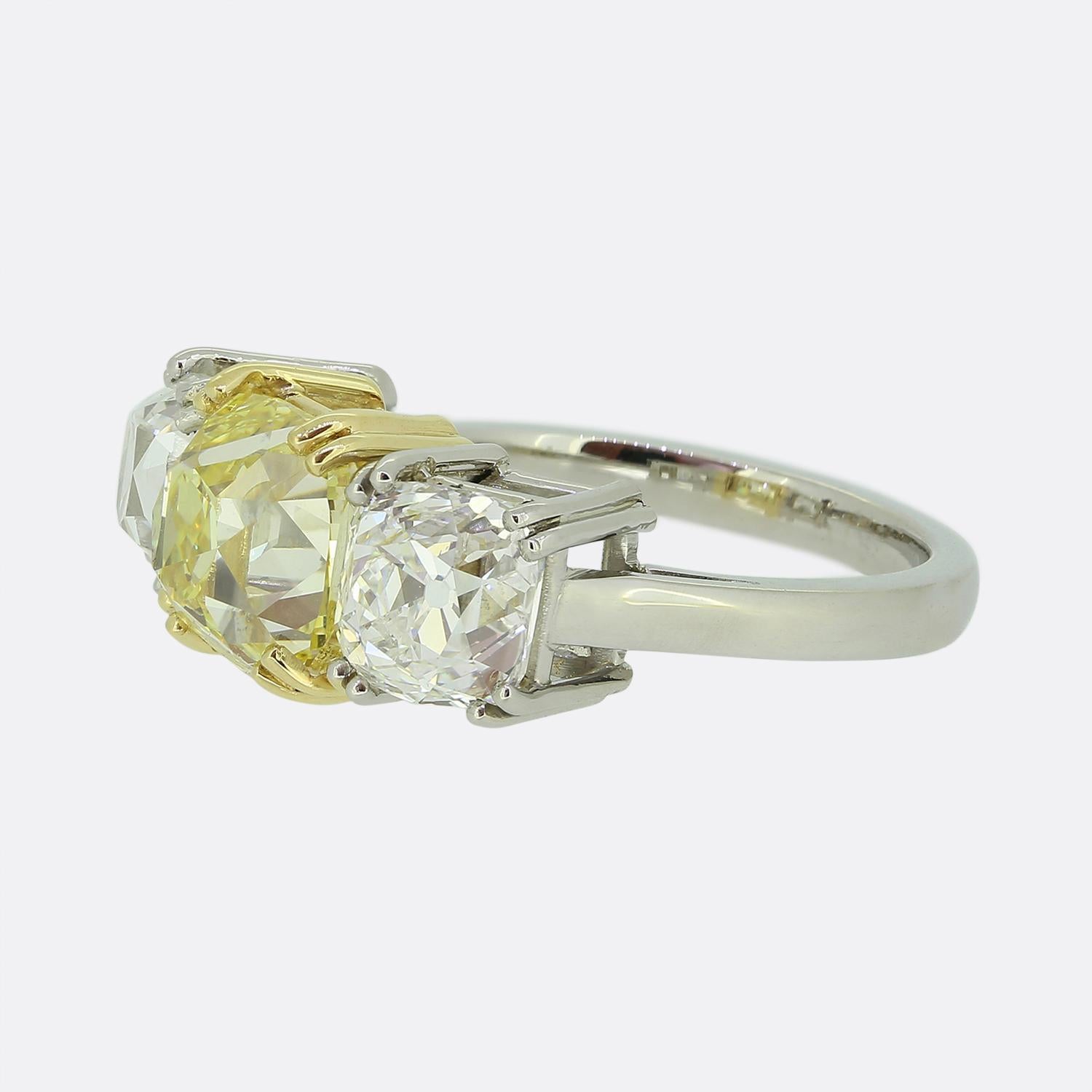 Here we have an exceptional three-stone diamond ring. At the centre of the face we find a breathtaking GIA certified old mine cut diamond possessing a natural fancy yellow colour tone  weighing an impressive 3.02 carats which sits in a rich yellow
