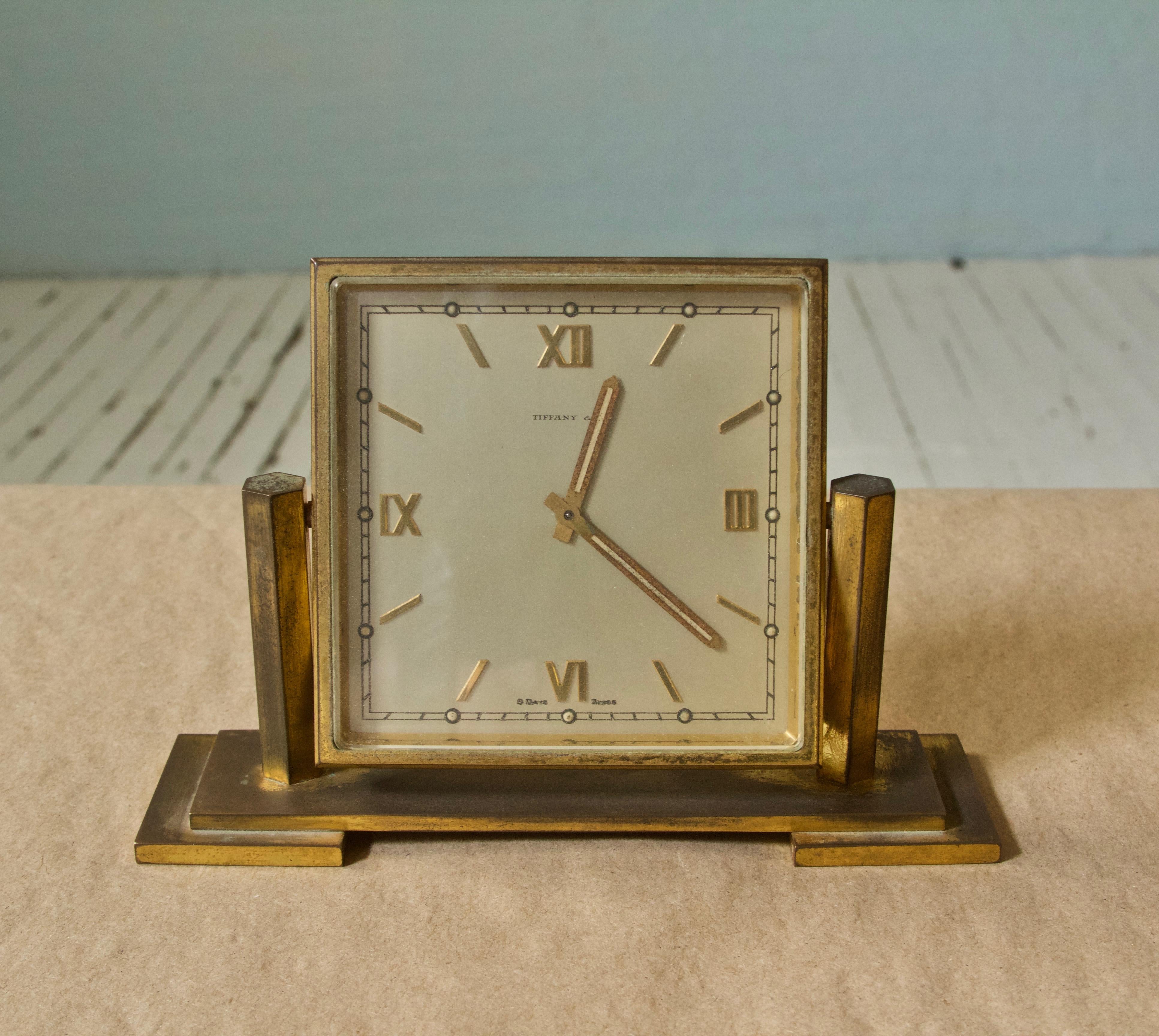 Handsome 8 day, Swiss jeweled Tiffany and Co. square desk clock in patinated brass with stepped dial frame and two-tiered base. Face pivots between flanking hexagonal columns for adjustable clock face angles. Two clock hands and applied Roman