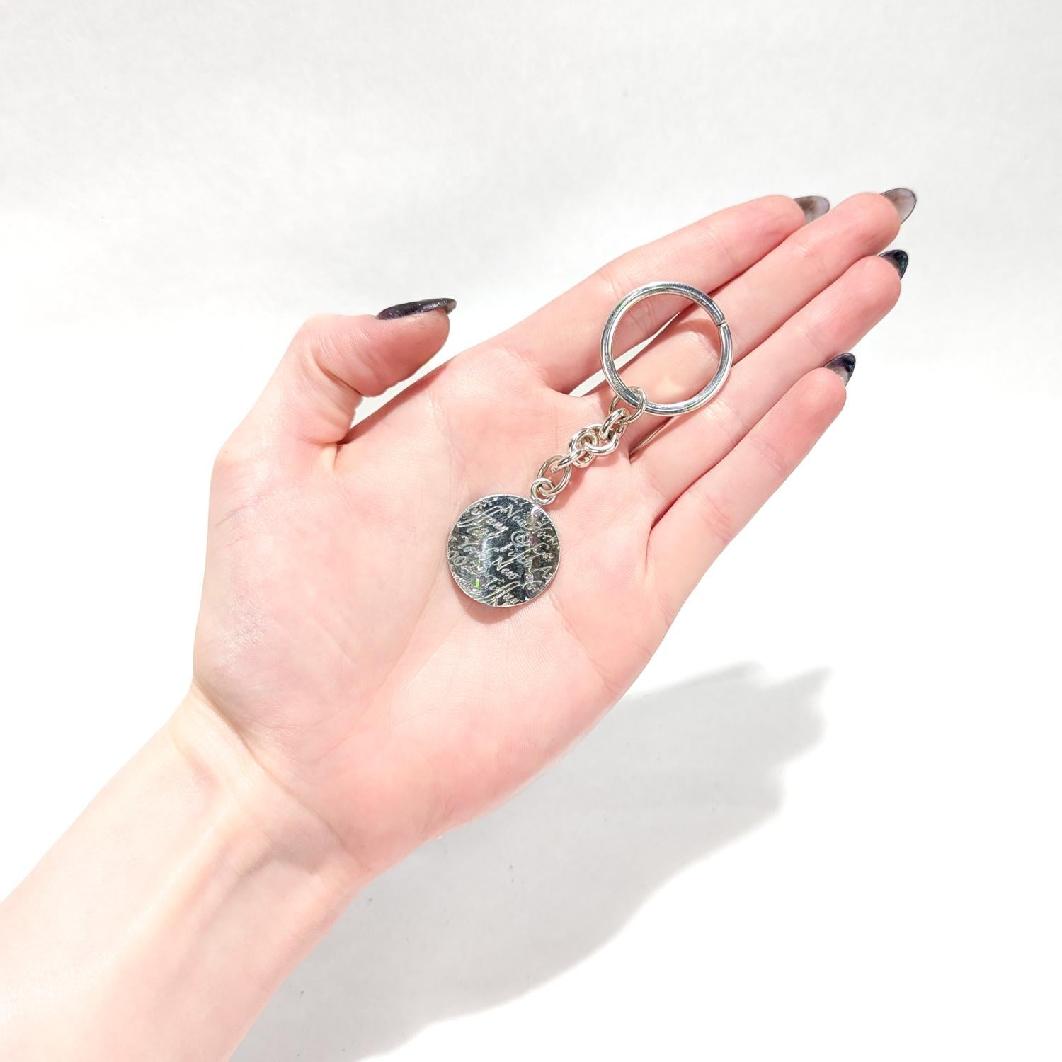 Un portachiavi in argento pregiato.

Da Tiffany & Co.

In argento sterling.

Con un portachiavi e un ciondolo collegati da una catena. L'etichetta è leggermente ondulata e decorata con una nuvola di parole con incise frasi in corsivo come 