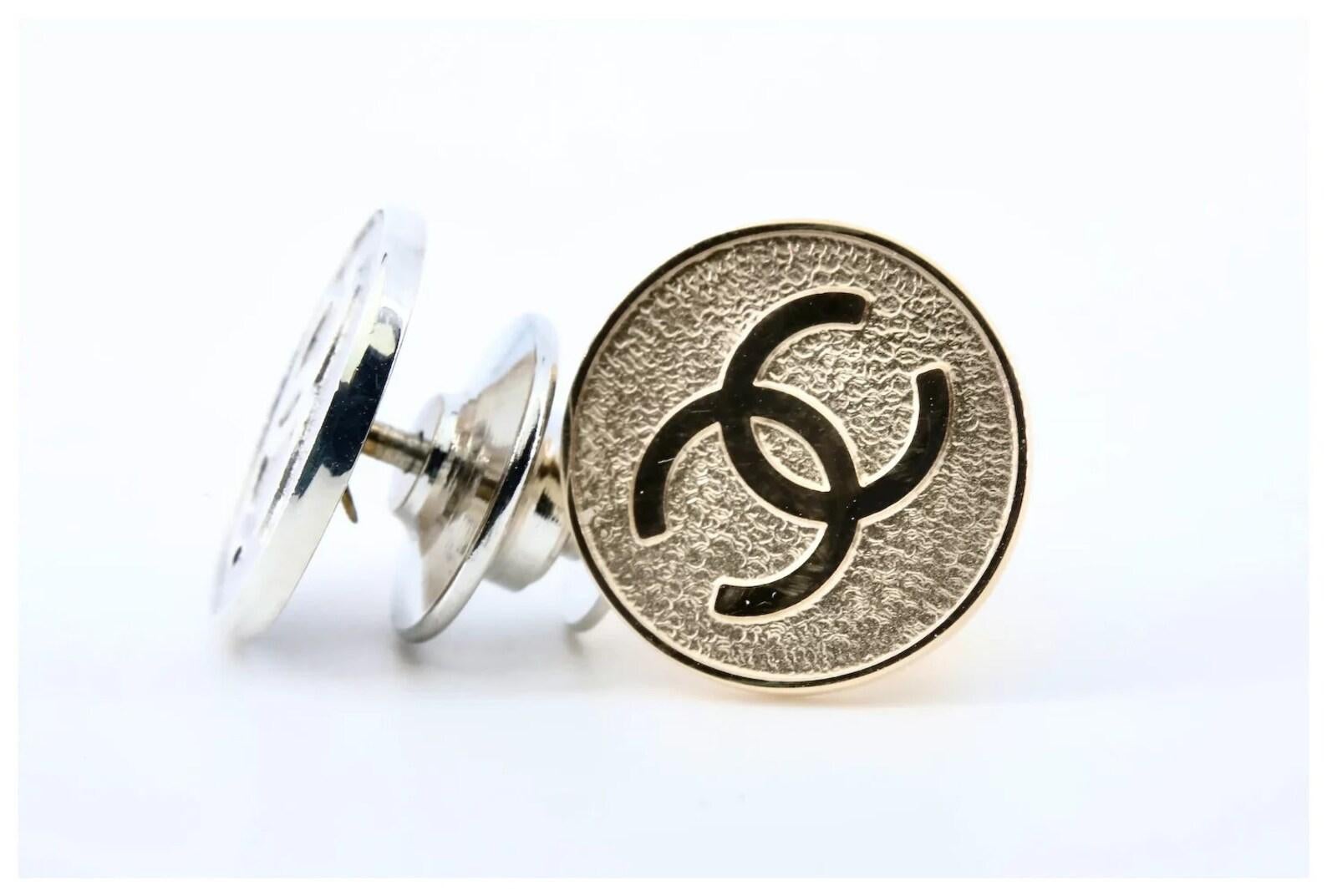 A pair of Tiffany & Company lapel pins for Chanel, one of 14 karat yellow gold and the other of sterling silver.

Displaying the raised polished Chanel logo in center framed by a matte background.

Stamped Tiffany & Co, 14K gold, and Sterling