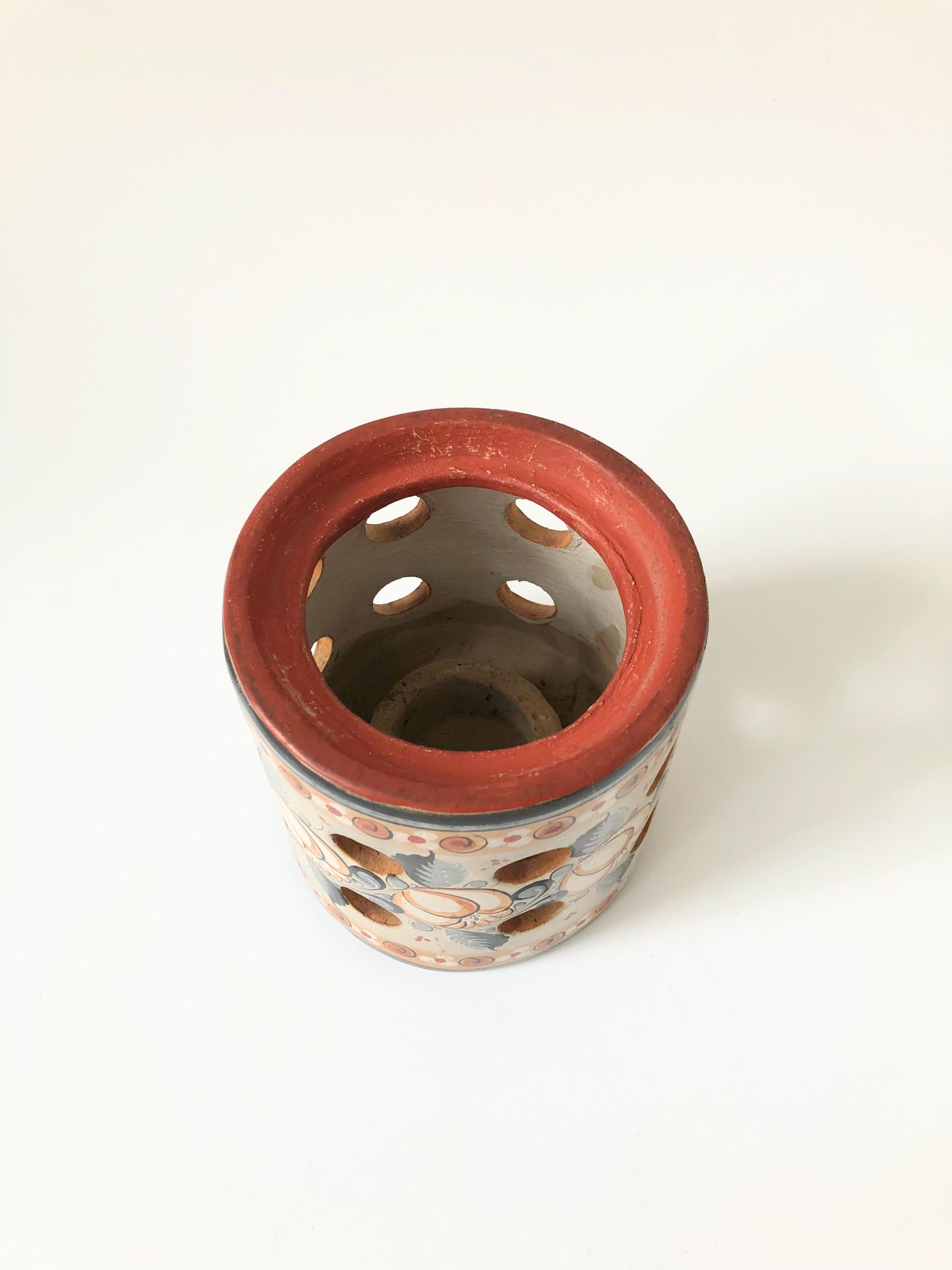 A vintage Mexican Folk Art tonala pottery candle holder. Beautiful hand painted designs wrap around the sides with circular cutouts for letting out the light. A circle is formed in the center for placing a votive candle.
 