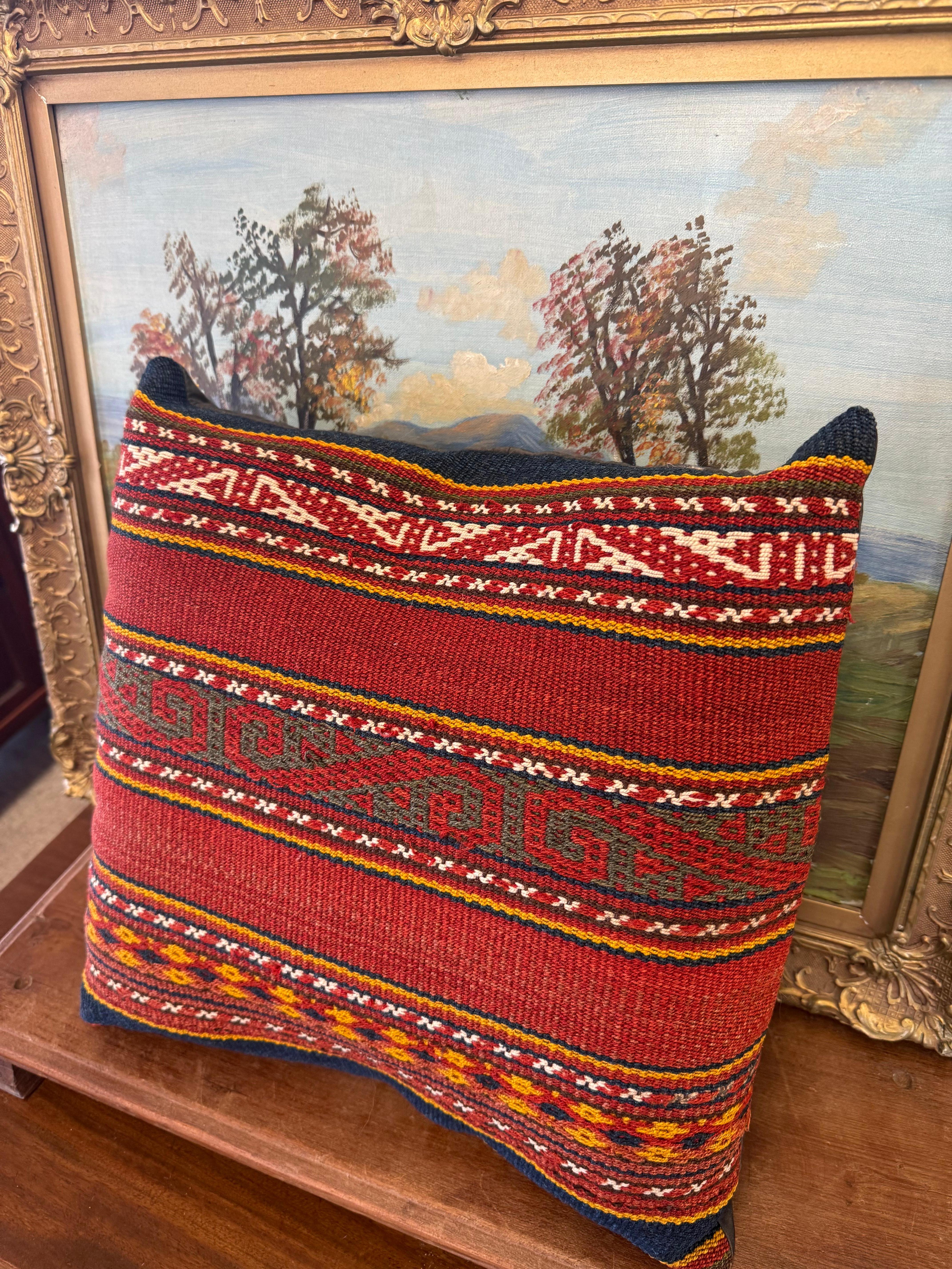 El cojín presenta un diseño tradicional tejido a mano con motivos geométricos y colores vivos como el rojo, el amarillo, el blanco y el azul. Este estilo es característico de los tejidos folclóricos o étnicos, a menudo asociados con culturas como