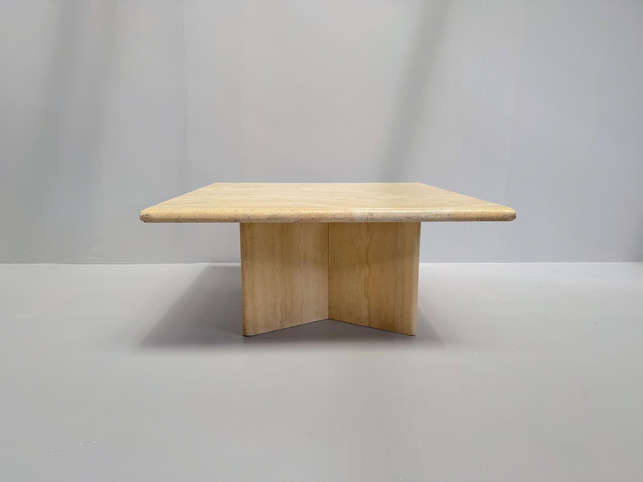Beautiful vintage travertine coffee table with sculptural presence and a minimalist aesthetic. Crafted from solid travertine stone, this piece features a thick square top with rounded edges and a heavy X-shaped pedestal base. The natural stone