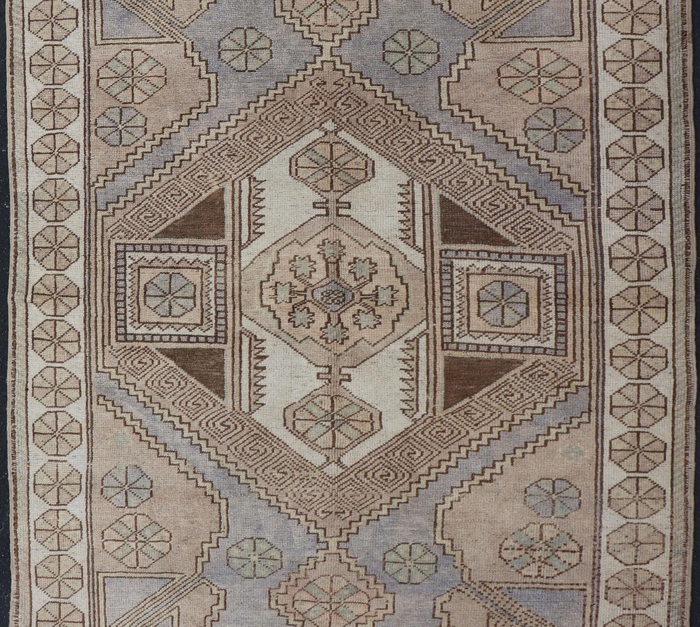 This vintage Turkish Oushak rug features a singular medallion design, filled with geometric motifs and flanked by complementary motifs in the central field and surrounding border. The design is rendered in a variety of lavender and taupe color