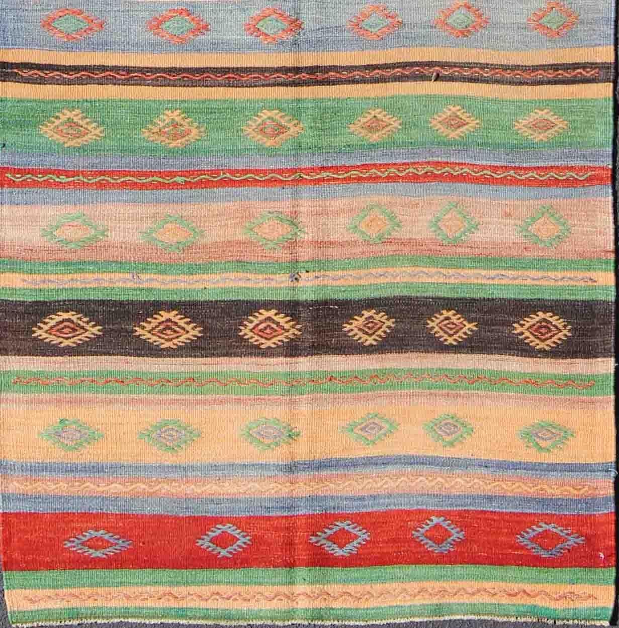 Featuring tribal shapes with a spotted and speckled assortment of geometric elements, rendered in a repeating stripe design, this unique Mid-Century Kilim showcases an array of colorful tones, including green, light blue, sand, charcoal and