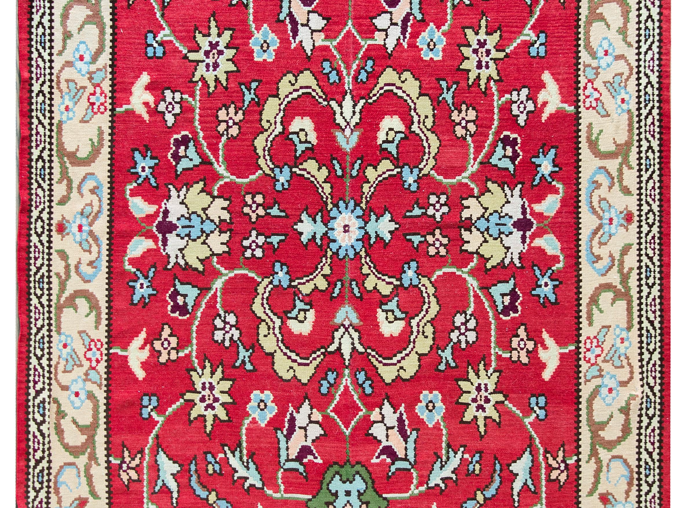A vintage Turkish kilim rug with a wonderful mirrored floral pattern woven in light and dark indigo, green, gold and brown, set against a bold crimson background, and surrounded by a beautiful scrolling vine and floral patterned border.
