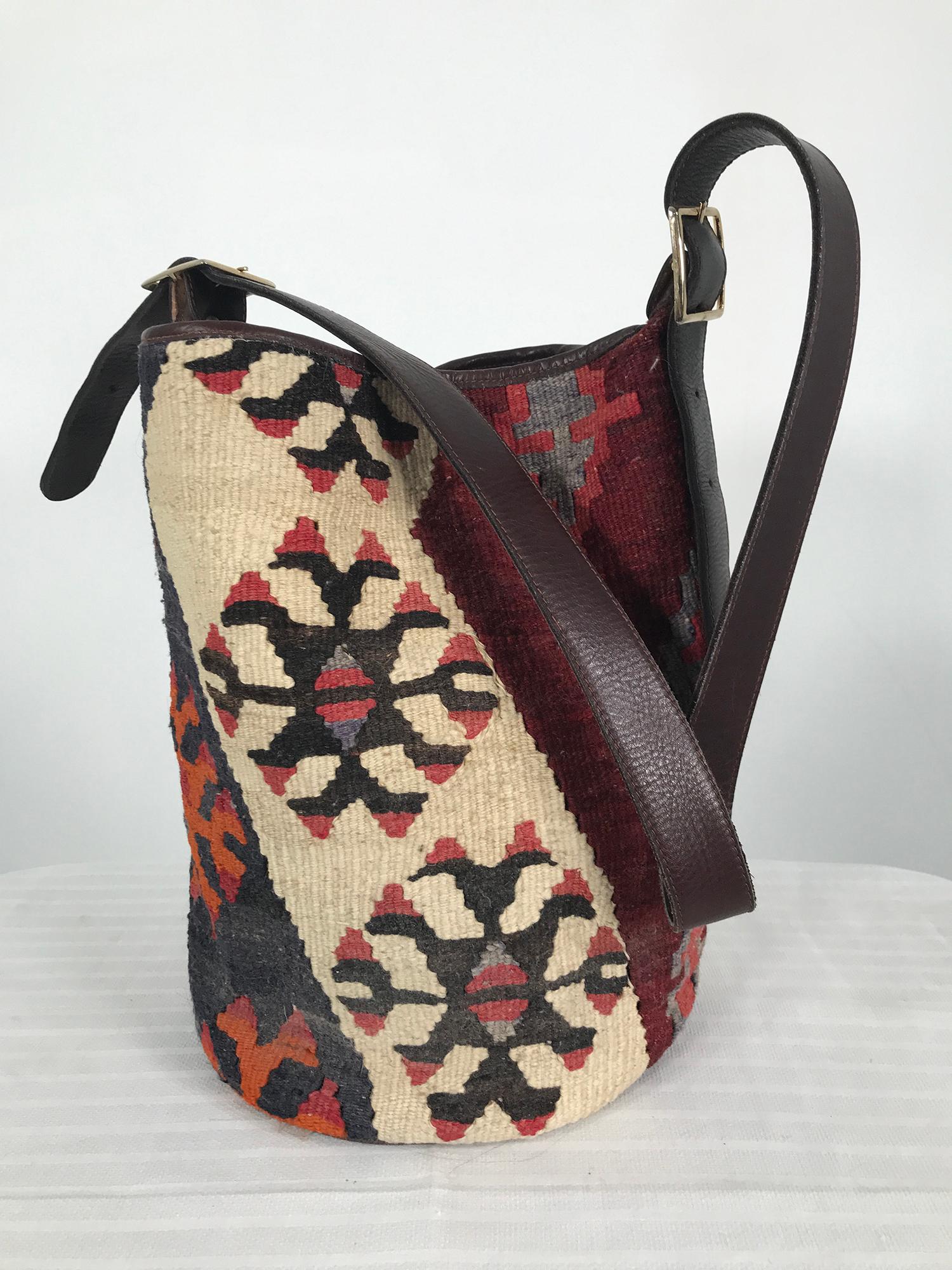 Vintage Turkish kilim rug with leather bucket shoulder bag. Woven wool kilim with leather trim, bottom and adjustable strap. The bag closes at the top with a zipper, it is lined in brown velvet like fabric, there is a single zipper compartment. The