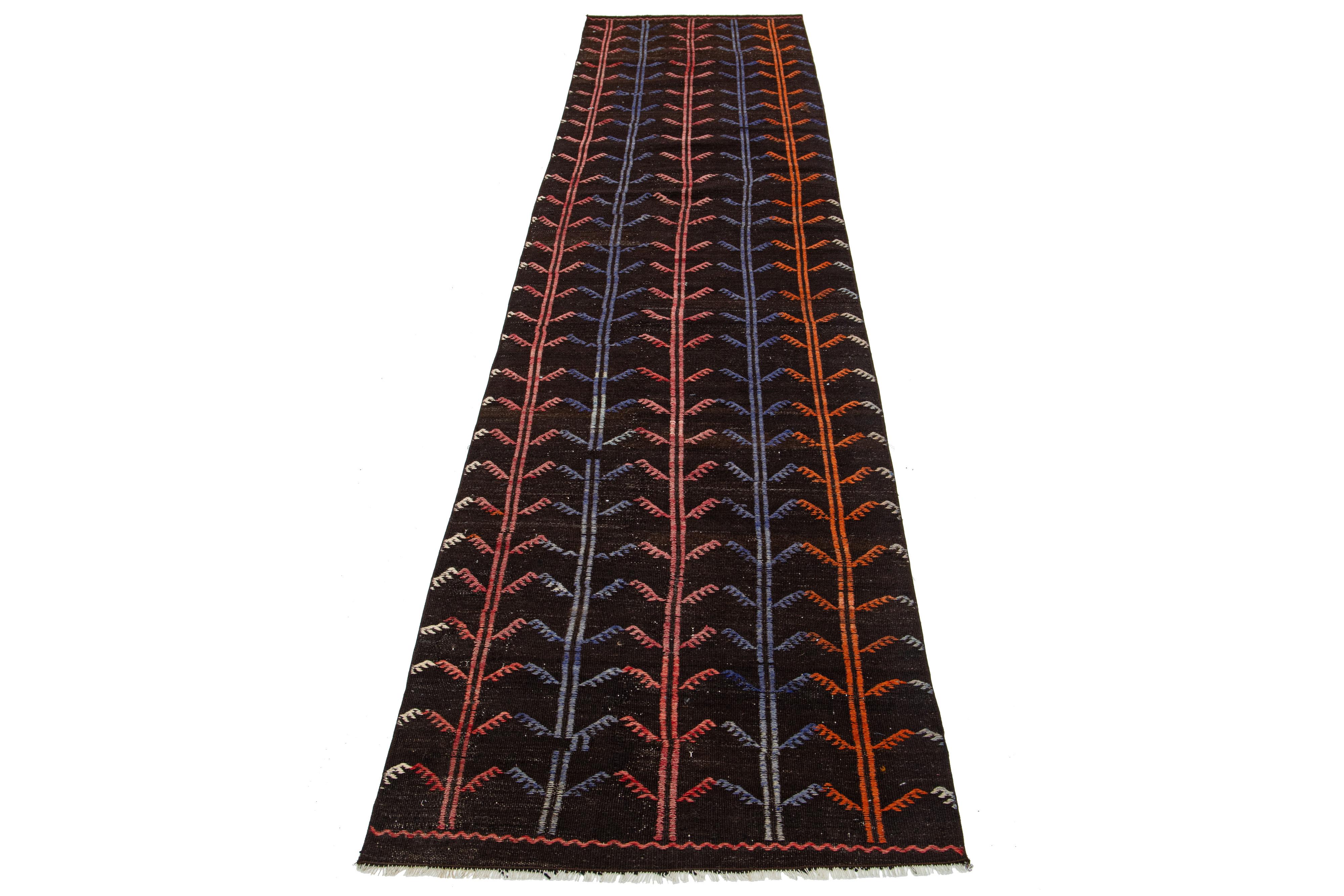 Ce magnifique tapis kilim en laine présente un étonnant design géométrique sur tout le pourtour, avec des accents multicolores vibrants sur un fond marron foncé.

Ce tapis mesure 2'10