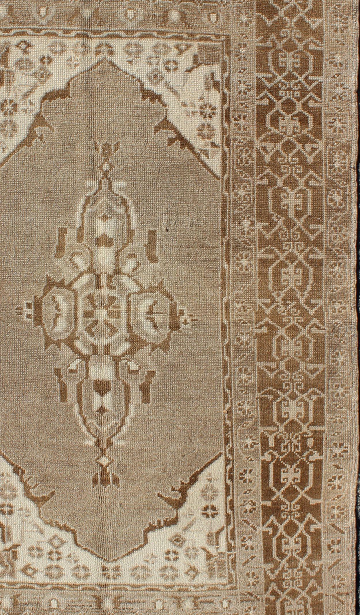 Das schöne und zarte Design dieses Teppichs mit seinem taupefarbenen und grauen Hintergrund und dem sanften Medaillon wird von sich wiederholenden geometrischen Elementen in Brauntönen eingerahmt. Die maskuline Bordüre verleiht dem Teppich ein