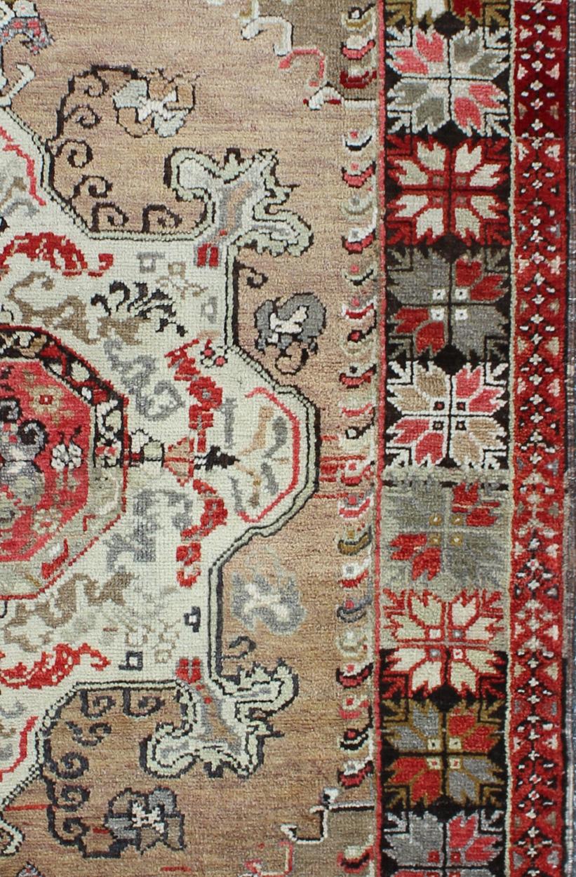 This vintage Turkish Oushak carpet (circa 1940) features a central blossoming medallion flower surrounded with smaller, expansive floral elements in the surrounding field. Colors include black and brown border, camel background with red, tan, taupe,