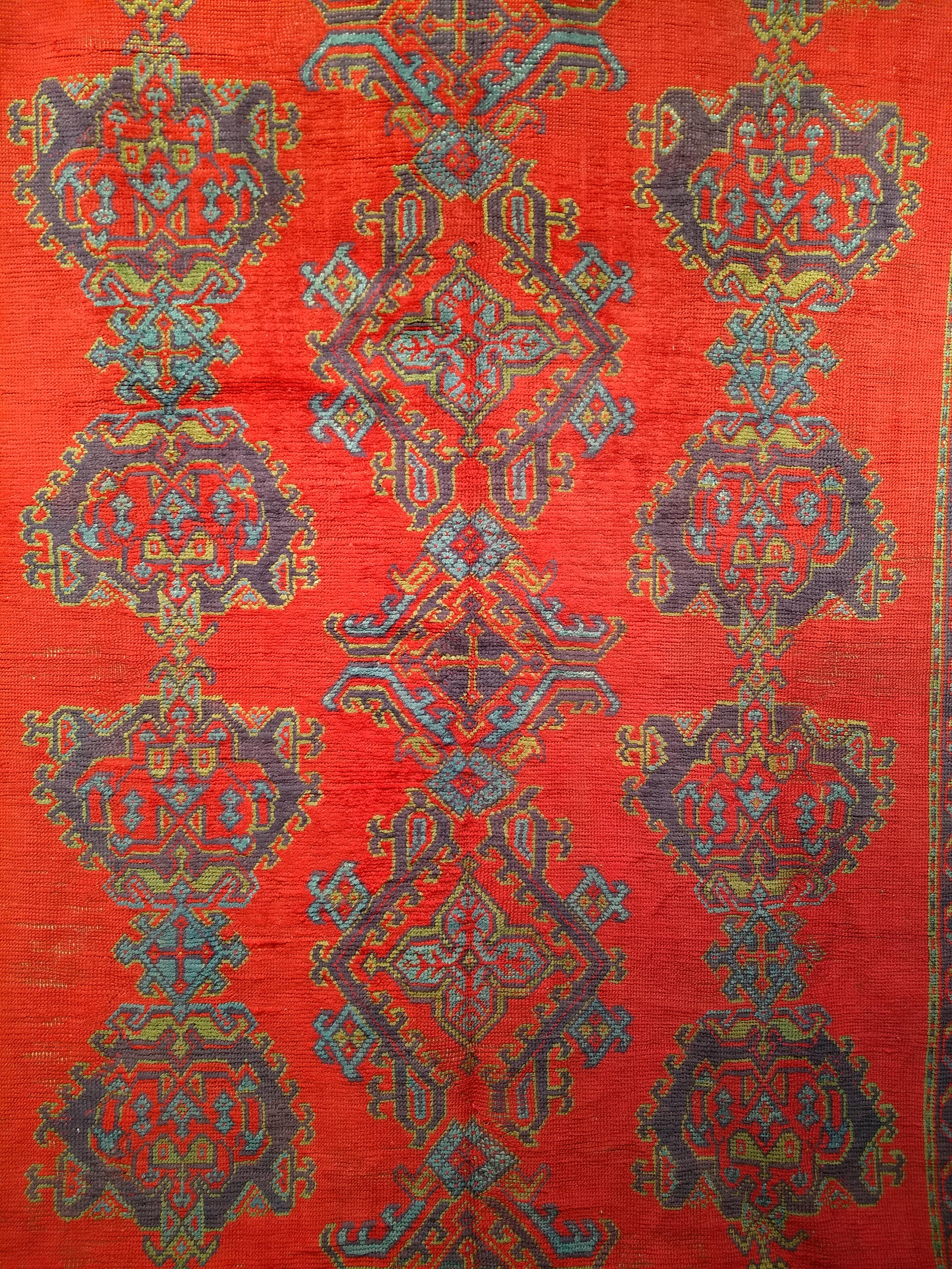 The  room size Turkish Oushak Rug has an all-over design set in a beautiful and rich red color hand-knotted in the early 1900s in Anatolia. There are three columns of large geometric design in colors of blue, green, purple, and yellow that are