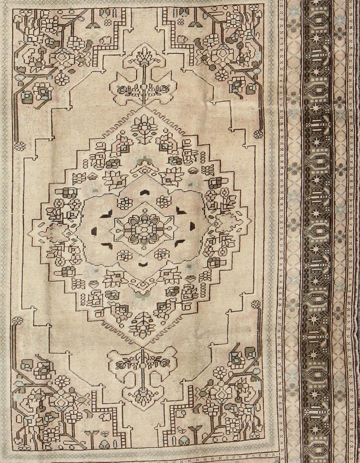 This striking vintage Turkish Oushak rug bears a neutral beige colored body that is highlighted by a dazzling design drawn in darker neutral earth tones. A large-scale medallion stretches across the central field. The encroaching multi-tiered border
