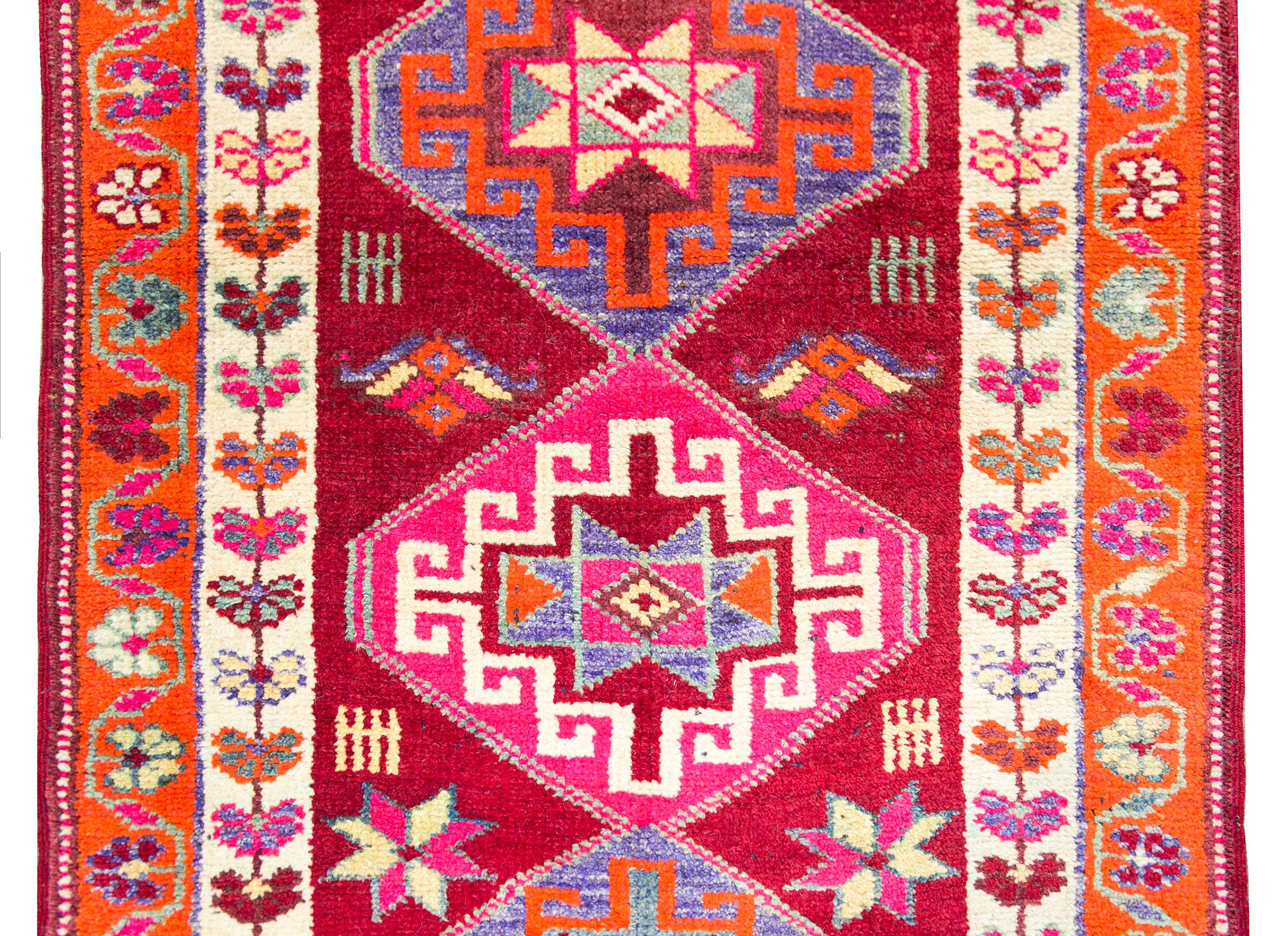 A stunning mid-20th century Turkish runner with multiple diamond medallions with geometric patterns woven in yellow, pink, orange, white, and indigo, and set against a dark red field with more stylized flowers, and surrounded by two floral patterned