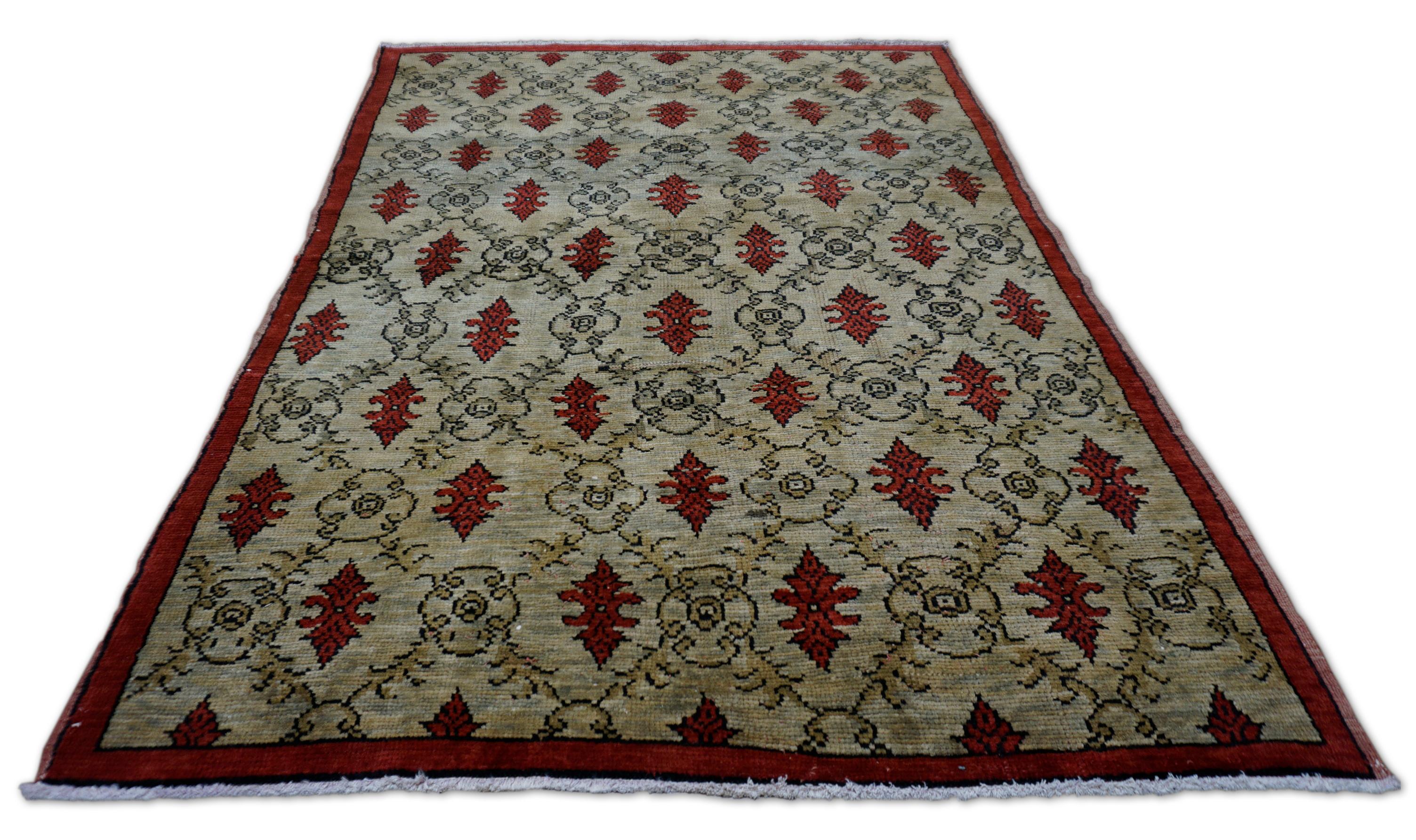 Dieser handgeknüpfte Wollteppich im Vintage-Stil besticht durch sein Zusammenspiel von rhythmischer Geometrie und traditionellen Motiven. Mit einem auffälligen All-Over-Rapport aus karminroten Blumenmedaillons, die in versetzten Reihen über einem