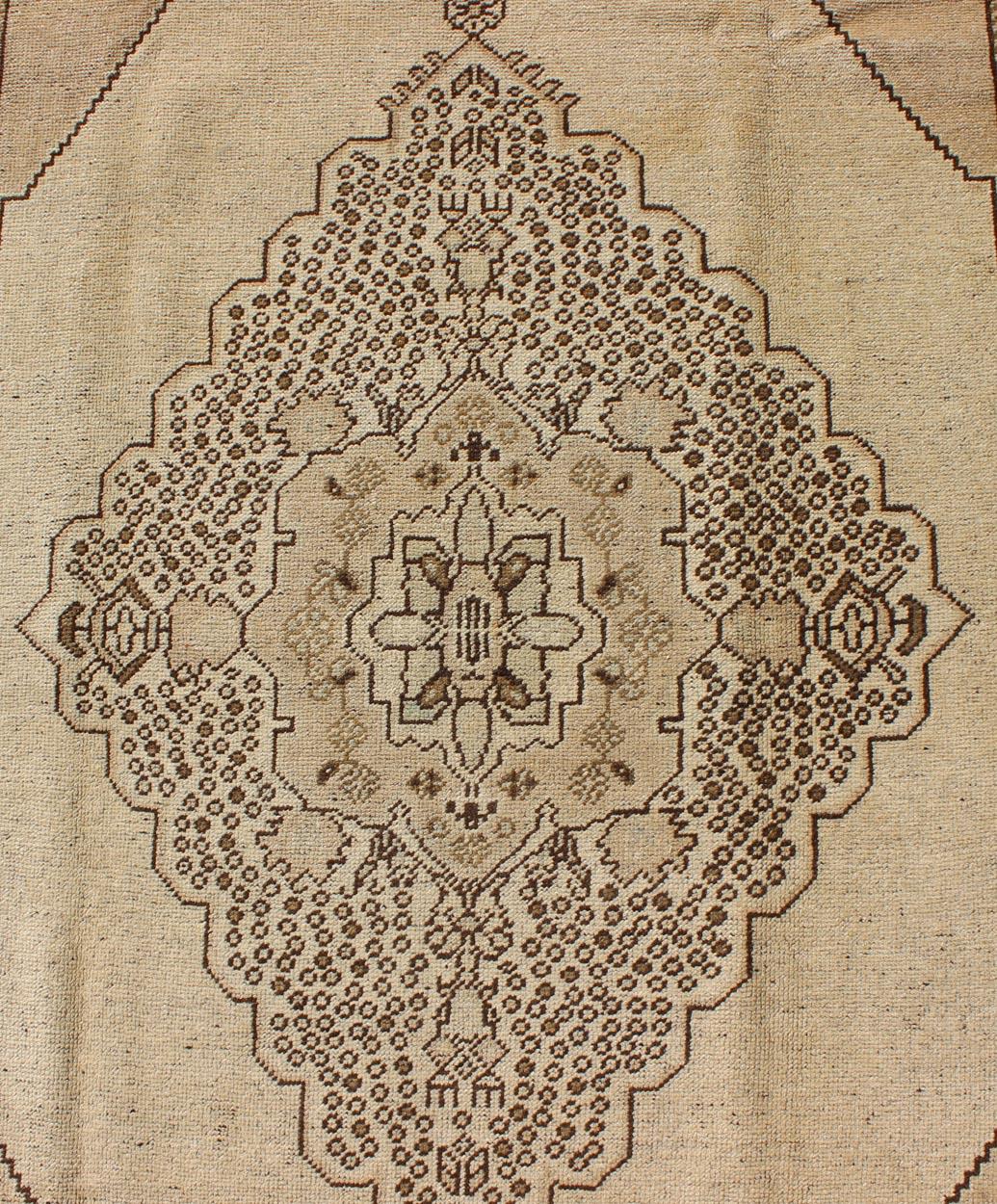 Measures: 5'7 x 10'2

This vintage Turkish Oushak sets on a neutral sandy ground with a central medallion stylized with small details. The background is pale khaki color, while the border has a botanical pattern of vines and flowers with light
