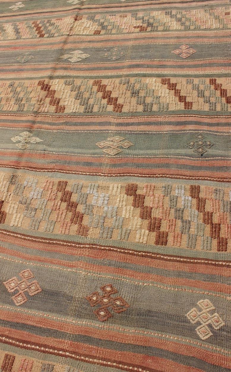 Measures: 5'4 x 9'8

This lovely Turkish Kilim has a striped, repeating pattern with clover motifs. The dark taupe-green stripes home the embroidered clovers, whereas the light clay colored stripes home geometric designs, reversing the palette of