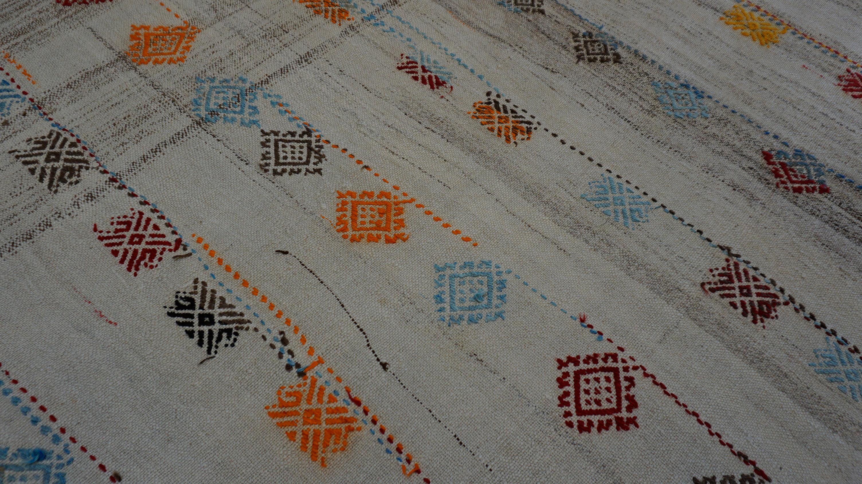 Este exclusivo kilim de lana tejida a mano presenta un cálido fondo marfil adornado con vibrantes motivos tribales dispuestos en una composición lúdica pero equilibrada. Filas de símbolos geométricos en forma de diamante -tejidos en rojo, naranja,