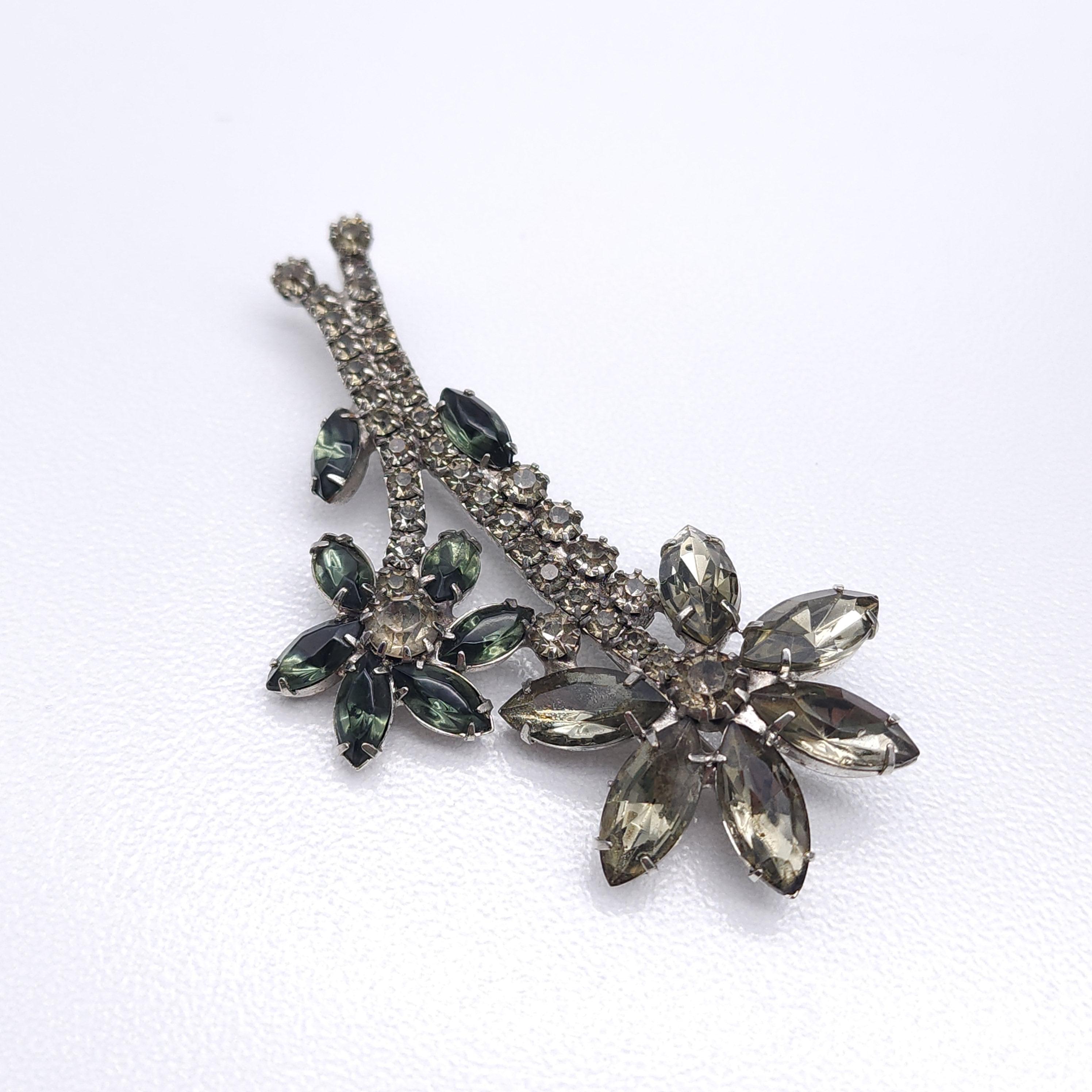 Dimensions: 3 inches x 1.5 inches
Marks/Hallmarks: n/a
Materials: Silver tone metal, prong-set crystals

This elegant vintage brooch features a floral design adorned with prong-set crystals in smoky gray and deep blue-green hues. The hypnotizing