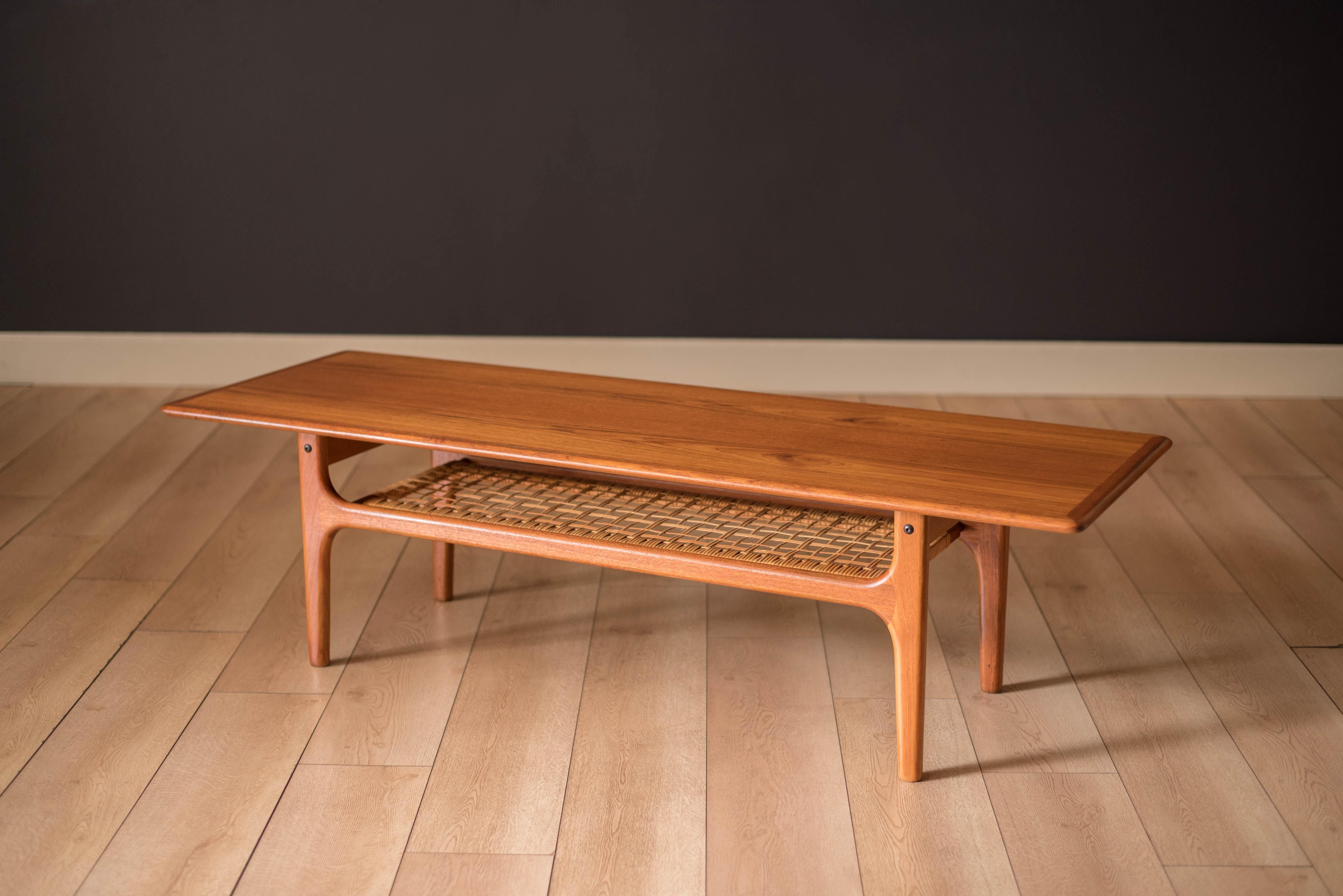 Mid century modern coffee table manufactured by Trioh, Denmark. This piece features sculpted edge banding and natural teak wood grains. Includes a lower tier magazine shelf for extra storage display made of hand woven cane kept intact.