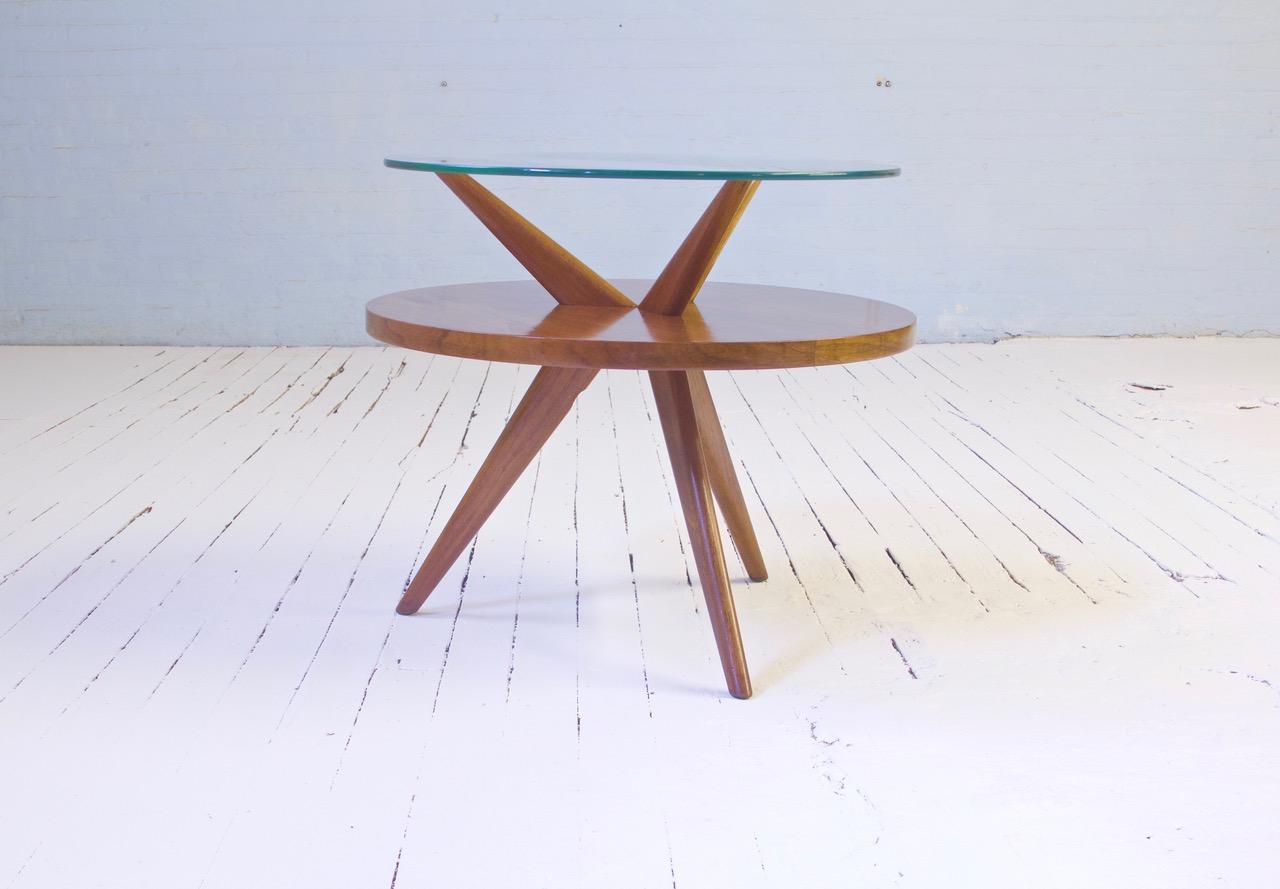 A well-preserved and dynamically-designed American mid-century two-tiered side table in Black Walnut and glass. Lovely lines throughout with sharply-splayed legs mirroring the upper supports creating a simple yet sophisticated visual dialogue which