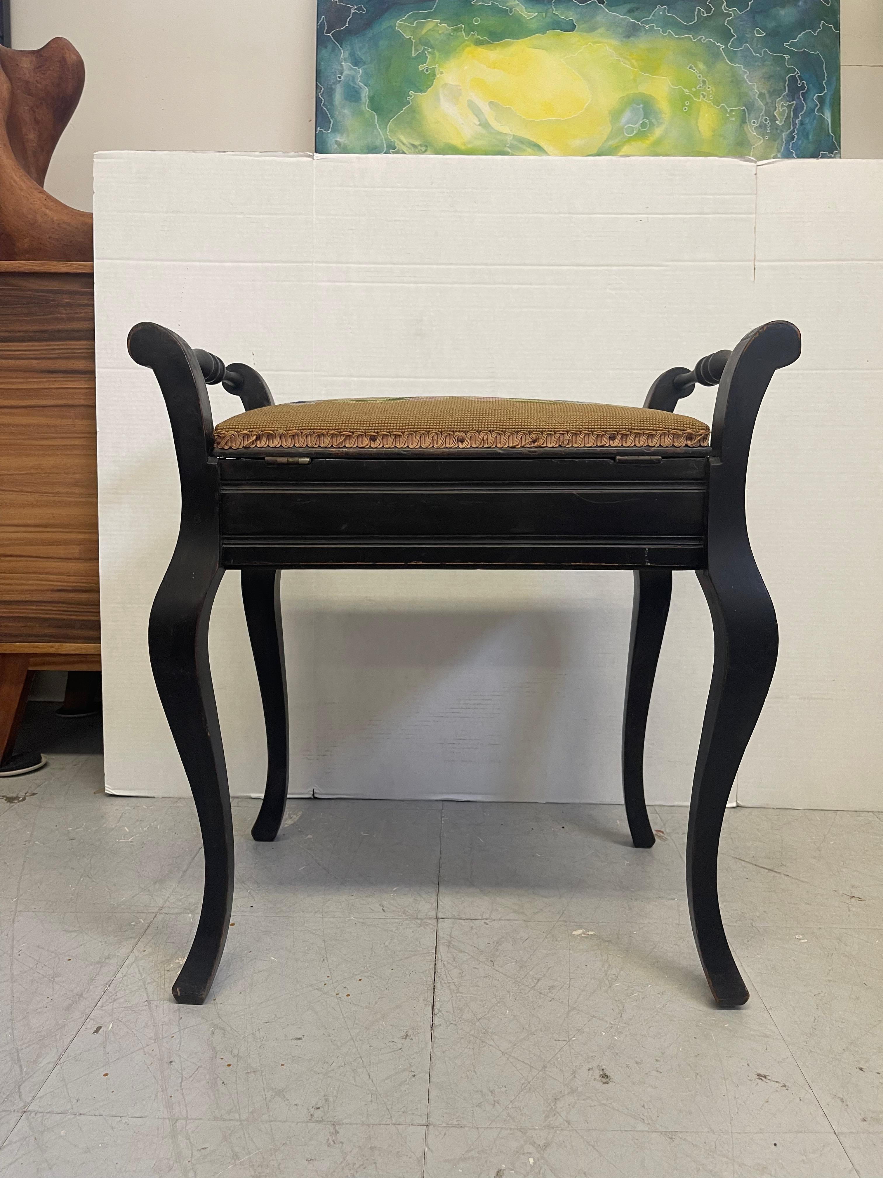  Vintage black toned wooden stool with curved cabriole legs and spindle side rails. The cushioned seat features a traditional floral needlepoint tapestry, framed with gimp trim. Imported from the UK, this compact bench works well as a vanity stool,