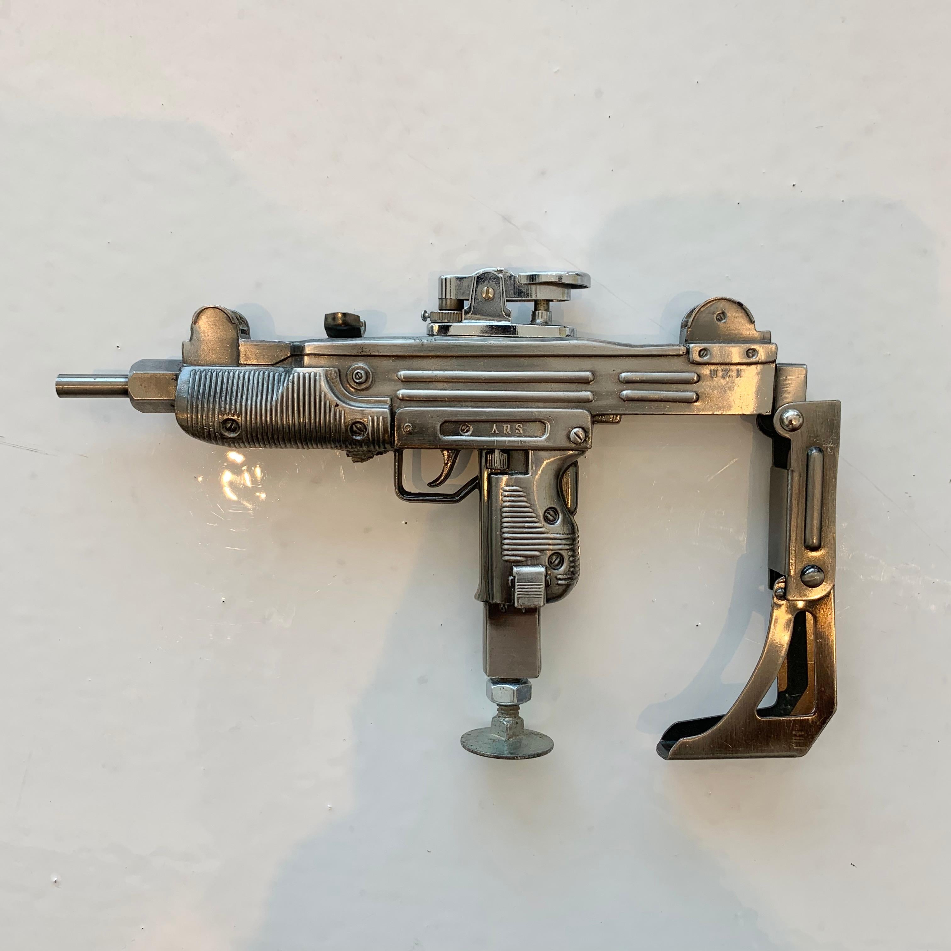 Cool vintage table lighter in the shape of an Uzi, Israeli machine gun. Made of metal and stands up on its own. Arm brace folds up and down. Cool tobacco accessory and conversation piece. Working lighter. 

  