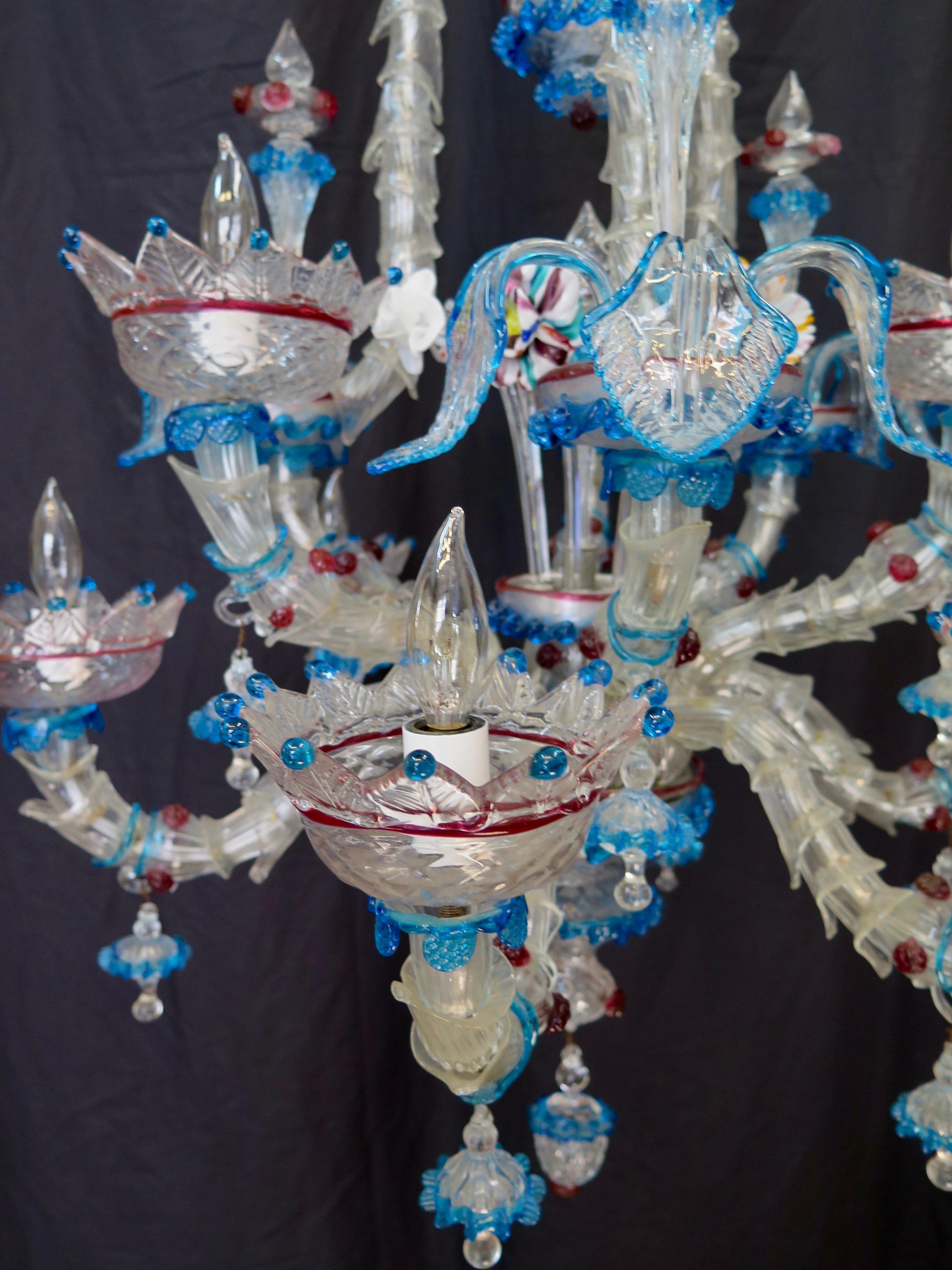 This elaborate midcentury Venetian glass chandelier is handmade by Murano. It is an exquisite double tier nine arm lighting fixture with hand blown embellishments of foliage accents and colorful flowers that accentuate its beauty. It will make a