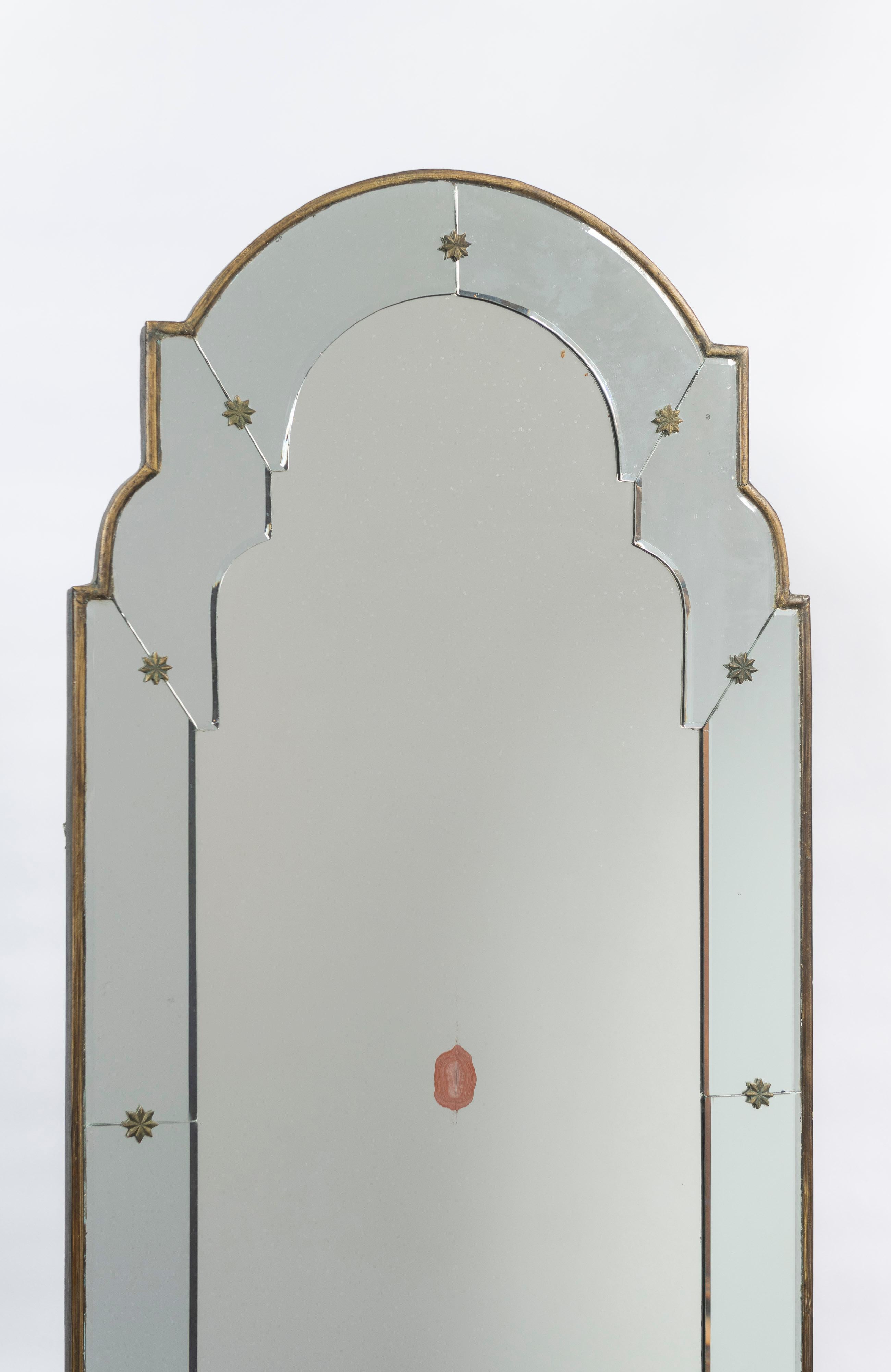 Le miroir mural de style vénitien est arqué avec un cadre biseauté, des formes florales en bronze laqué et un bord en bois doré. Le miroir présente plusieurs fissures visibles sur les photos ainsi que quelques zones abîmées. Bonne pièce pour créer