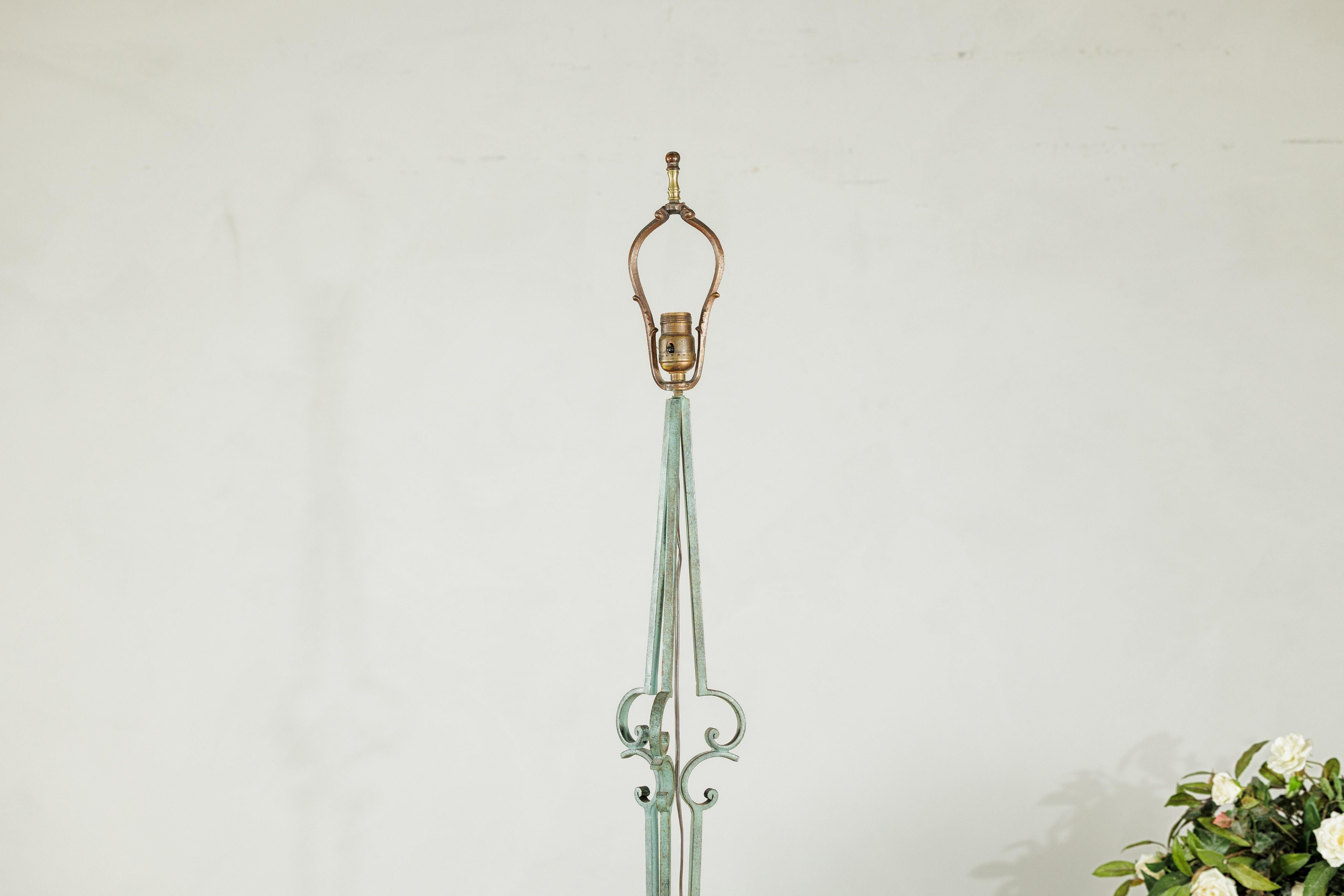 Questa lampada da terra vintage in ferro battuto presenta un'elegante forma allungata accentuata da una lavorazione a volute forgiata a mano e da una patina verdastra delicatamente invecchiata. Il design combina delicatezza e forza: sottili aste di