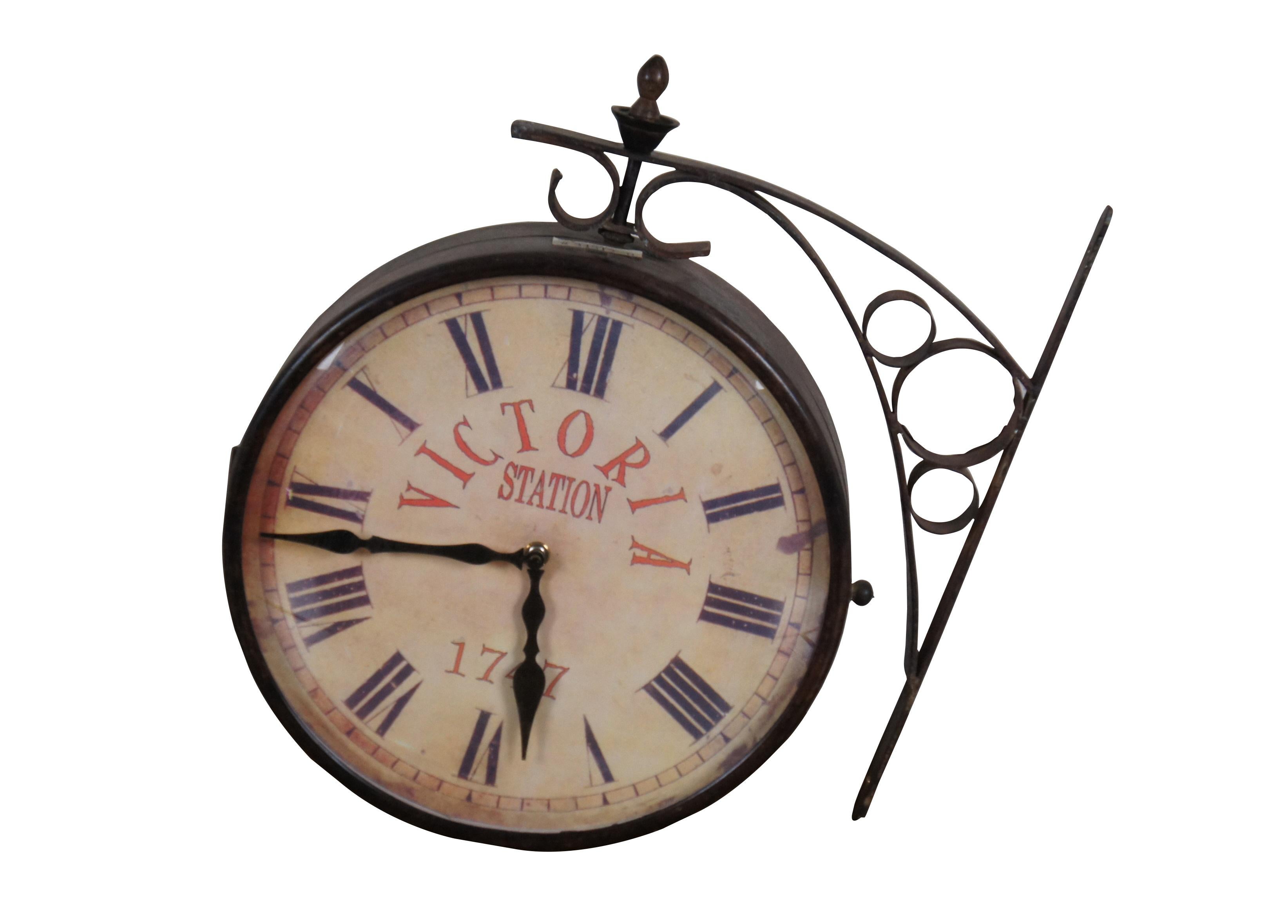 Late 20th century double sided railway clock styled after the famous clock at Victoria Station, London. Wrought metal wall mounted support and case in a dark bronze finish. 12 inch diameter printed face with faux distressing, Roman numerals, and the