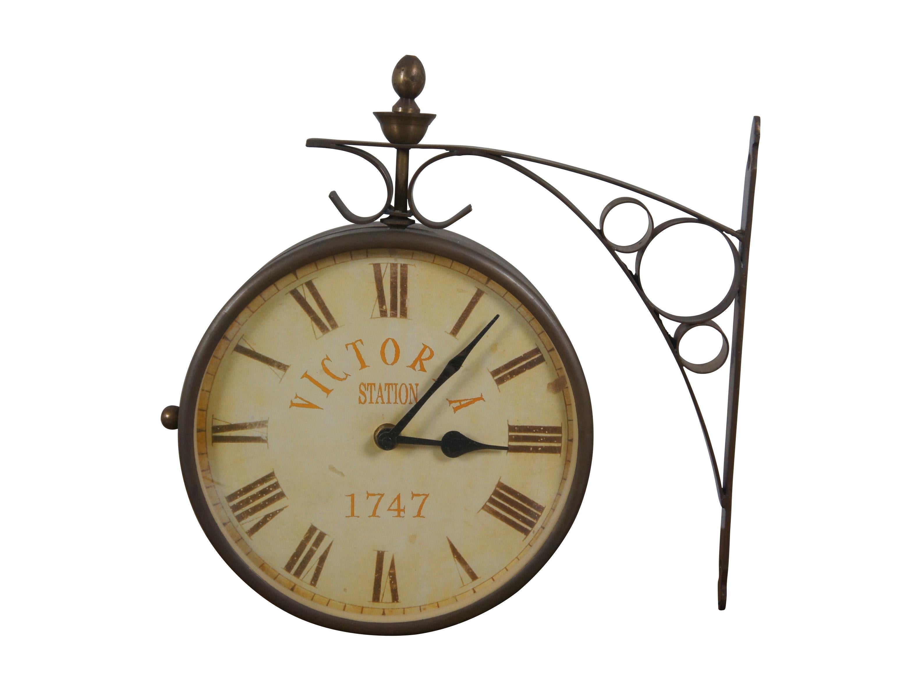 3 Available - Late 20th century double sided railway clock styled after the famous clock at Victoria Station, London. Wrought metal wall mounted support and case in a dark brass finish. 8 inch diameter printed face with faux distressing, Roman