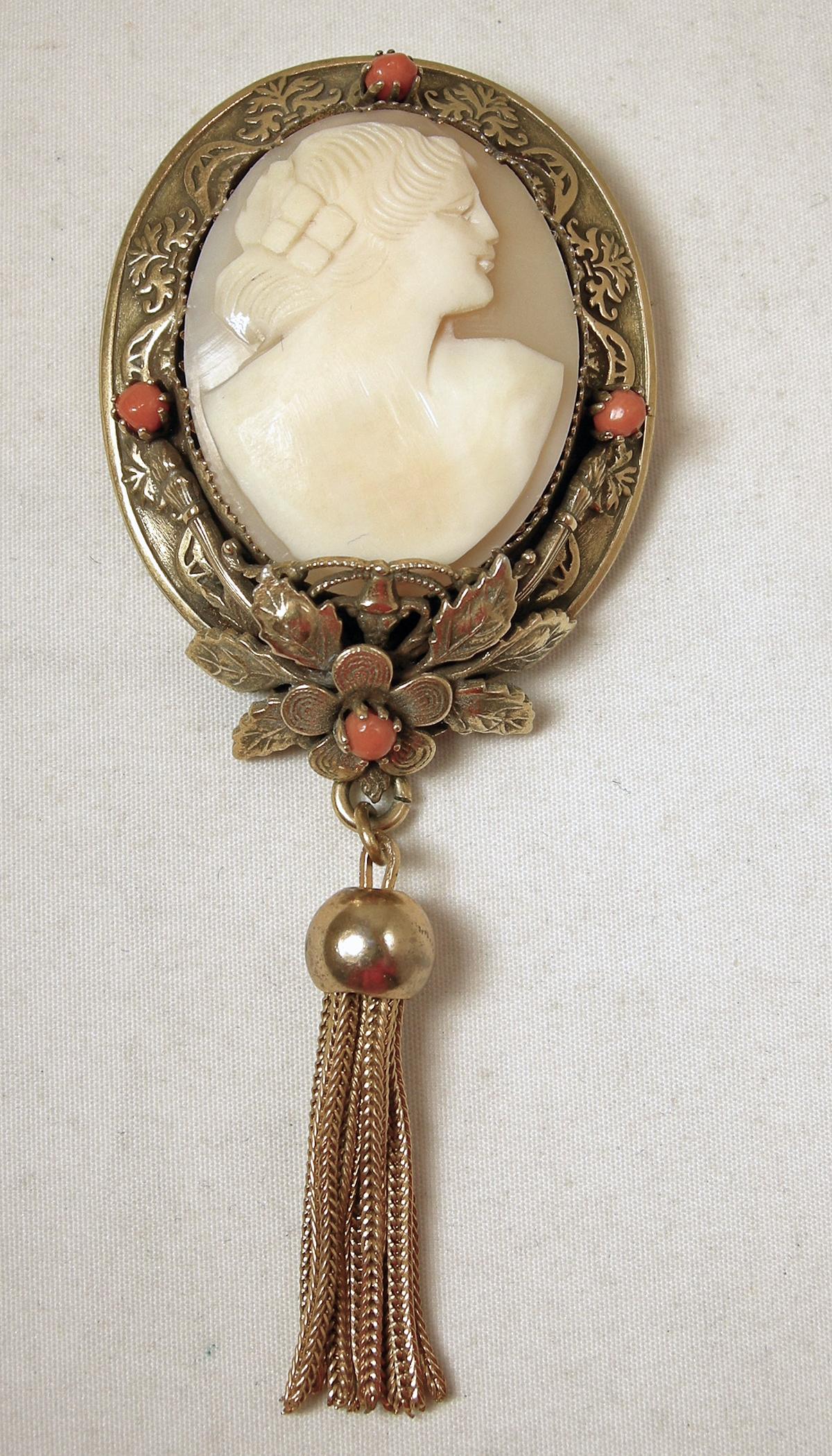 This vintage Victorian revival brooch can also be worn as a pendant. The brooch has a faux cameo center with faux coral accents on its gold tone border.  The brooch has tassels hanging downward. The brooch is 4” x 1-1/2” and the matching clip