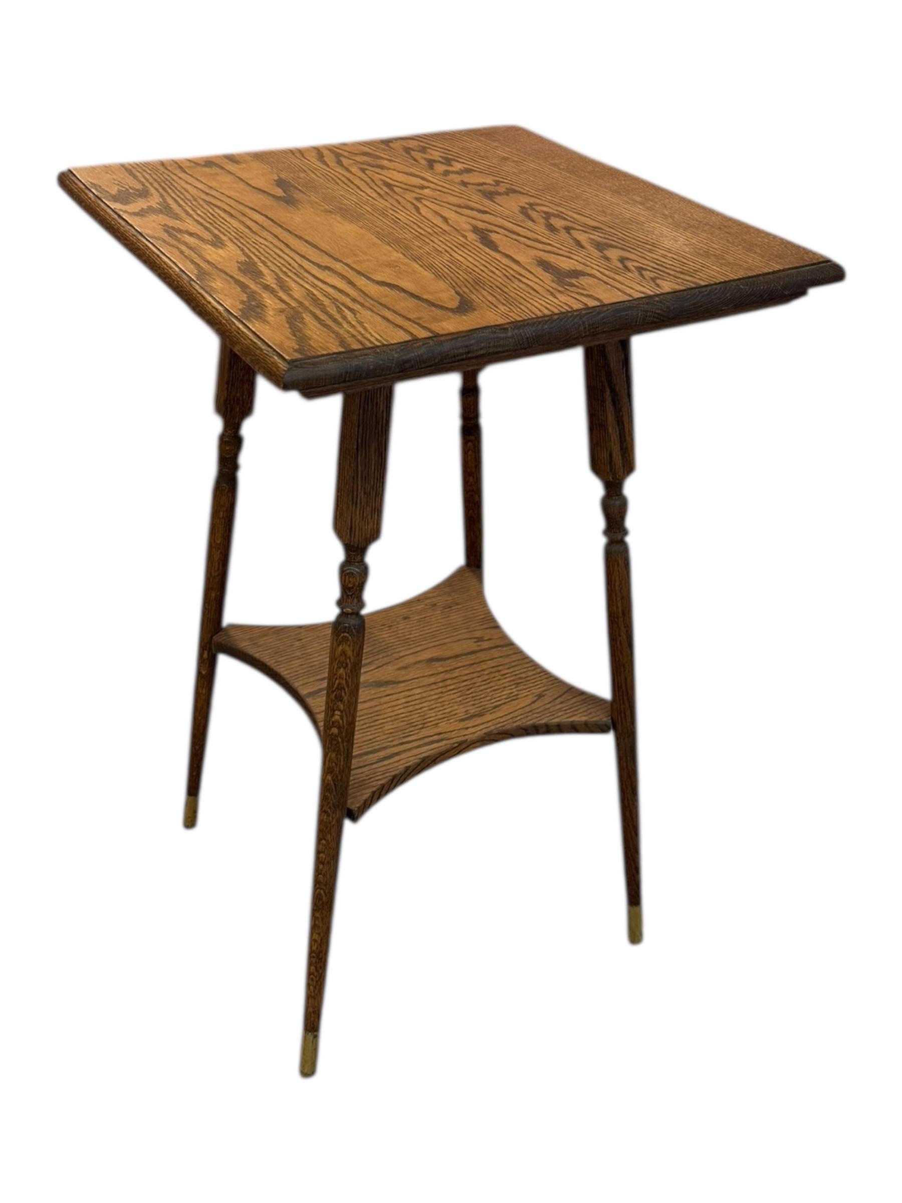  The table is a small, square, two-tiered side table made of oak with a rich, natural grain finish. It features four slender, tapered legs with turned detailing and brass-capped feet. The lower tier is a curved, open shelf, adding both functionality