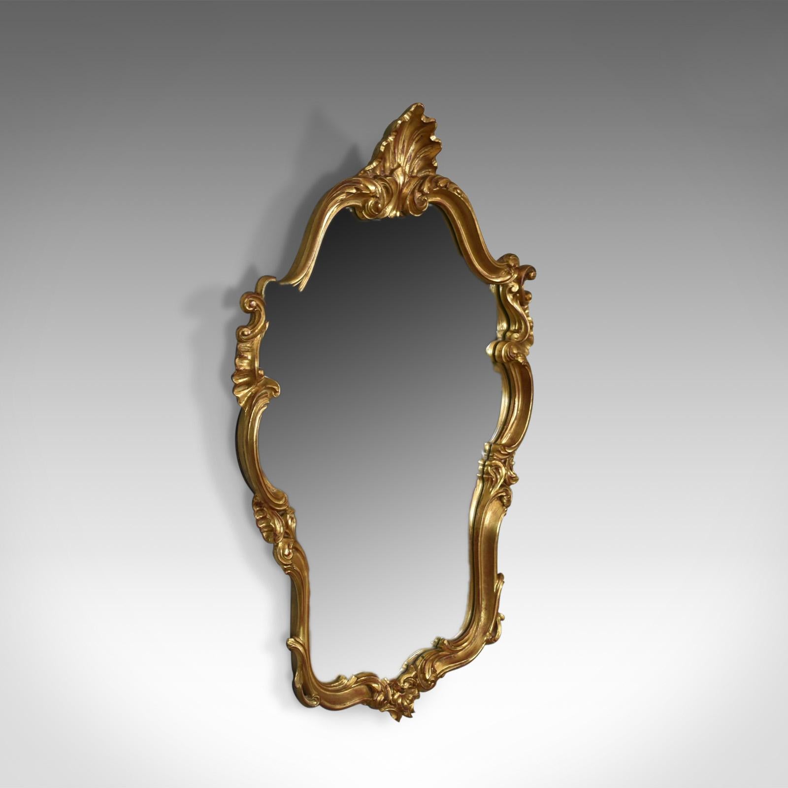 This is a vintage wall mirror in the Victorian Rococo Revival manner, English dating to the latter part of the 20th century.

Typically elaborate and elegant design
Giltwood frame featuring scrolls, leaves and embellishment
Quality shaped mirror