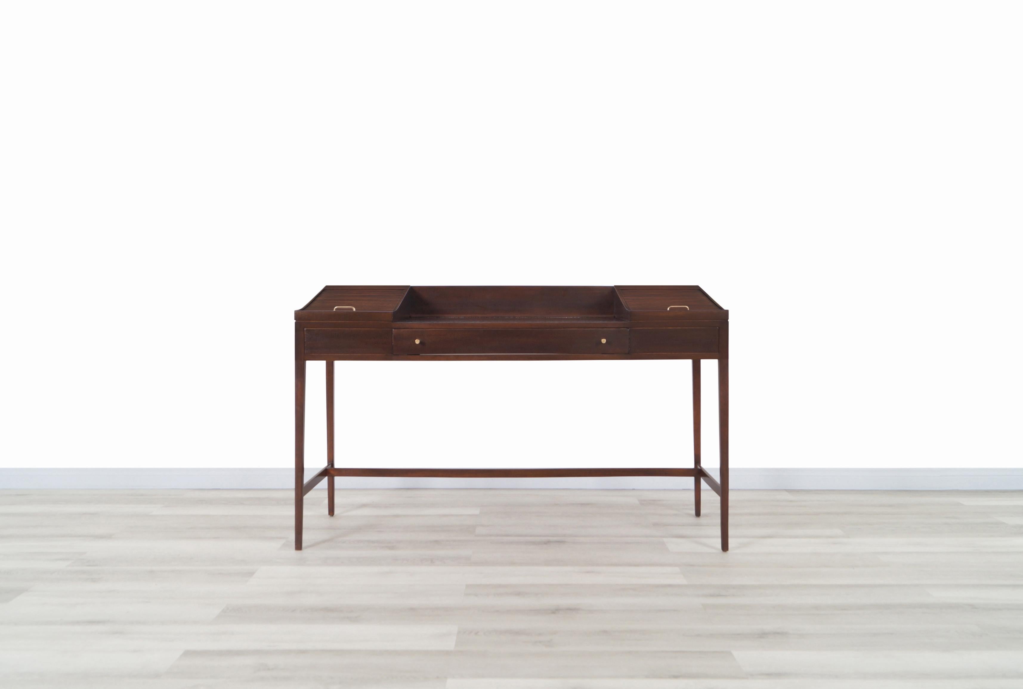 A beautiful vintage dark walnut desk manufactured in the United States, circa 1950s. Features two tambour doors that roll up, revealing one pull-out tray on each side, and the front is comprised of a large drawer for additional storage, all