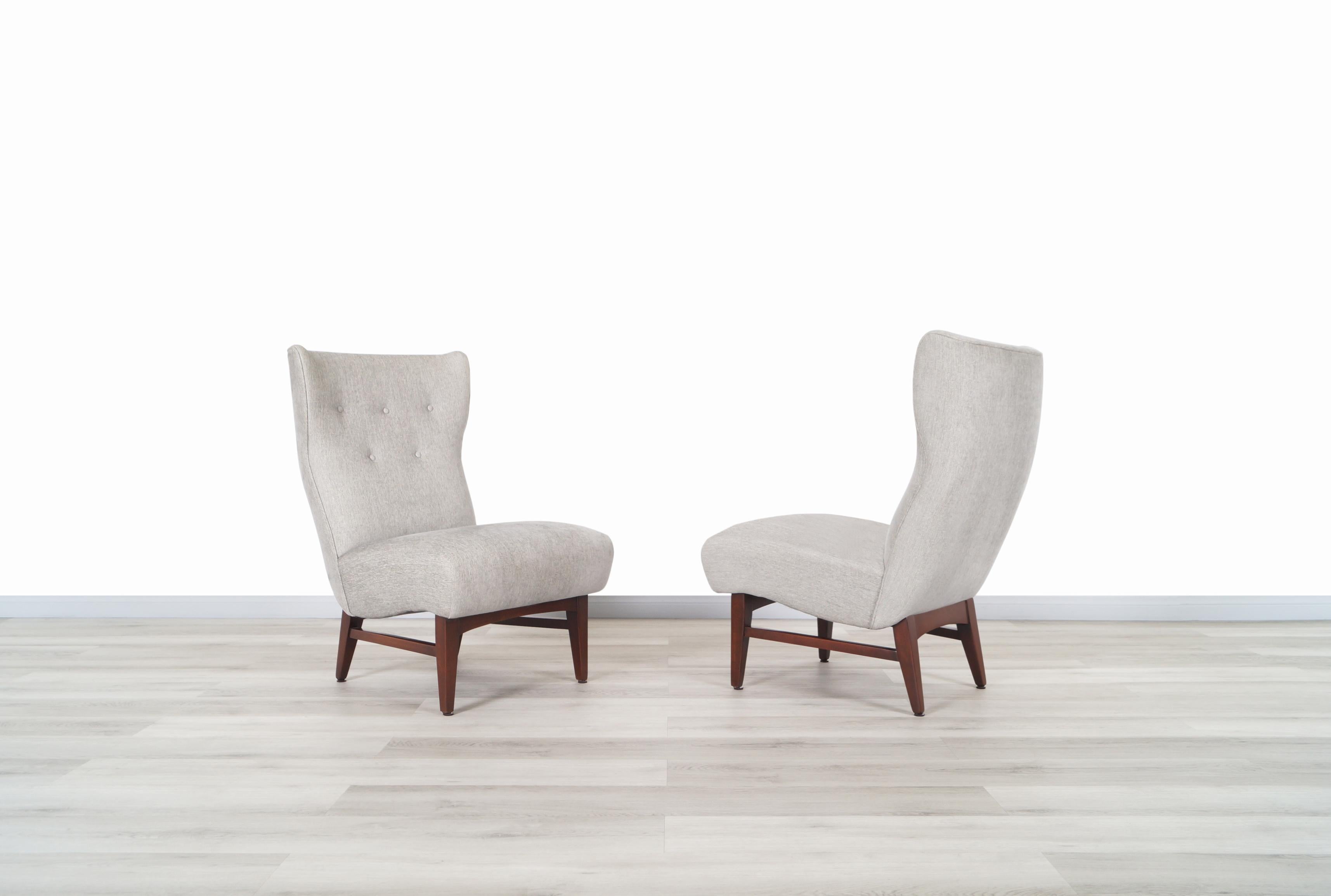 A beautiful pair of vintage wingback slipper lounge chairs manufactured in the United States, circa 1950s. These elegant chairs feature a sturdy solid walnut base and, newly upholstered seat in a shade of gray fabric with tufted details on the