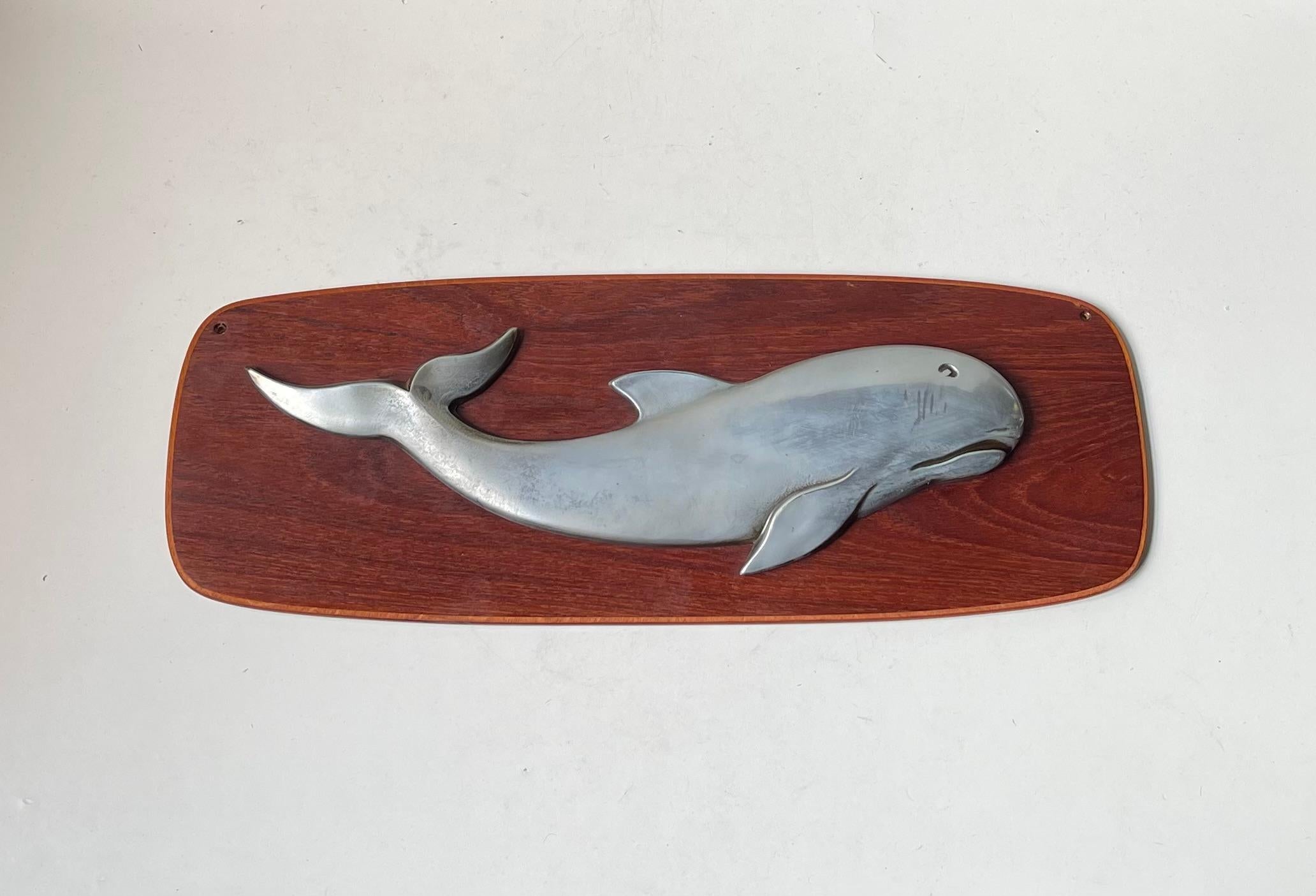 Eine stilvolle Wandtafel mit einem Wal, der auf einer Rückenplatte aus Teakholz schwimmt. Schöne 3-dimensionale Darstellung in Solid Aluminium. Seine sehr weichen Merkmale lassen vermuten, dass es sich um einen Schweinswal handeln könnte. Es wurde