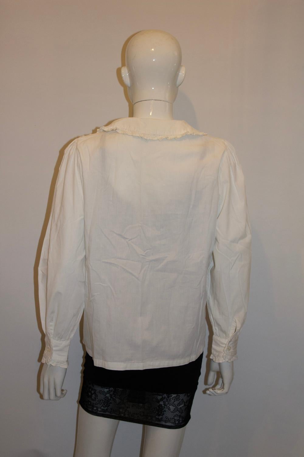 Un'ottima camicetta vintage per la primavera/estate. In cotone bianco di medio peso, la blusa ha un colletto a pan di Pietro con dettaglio di volant, maniche lunghe con polsino a un bottone e dettaglio di volant. Ha un'apertura frontale con bottoni,
