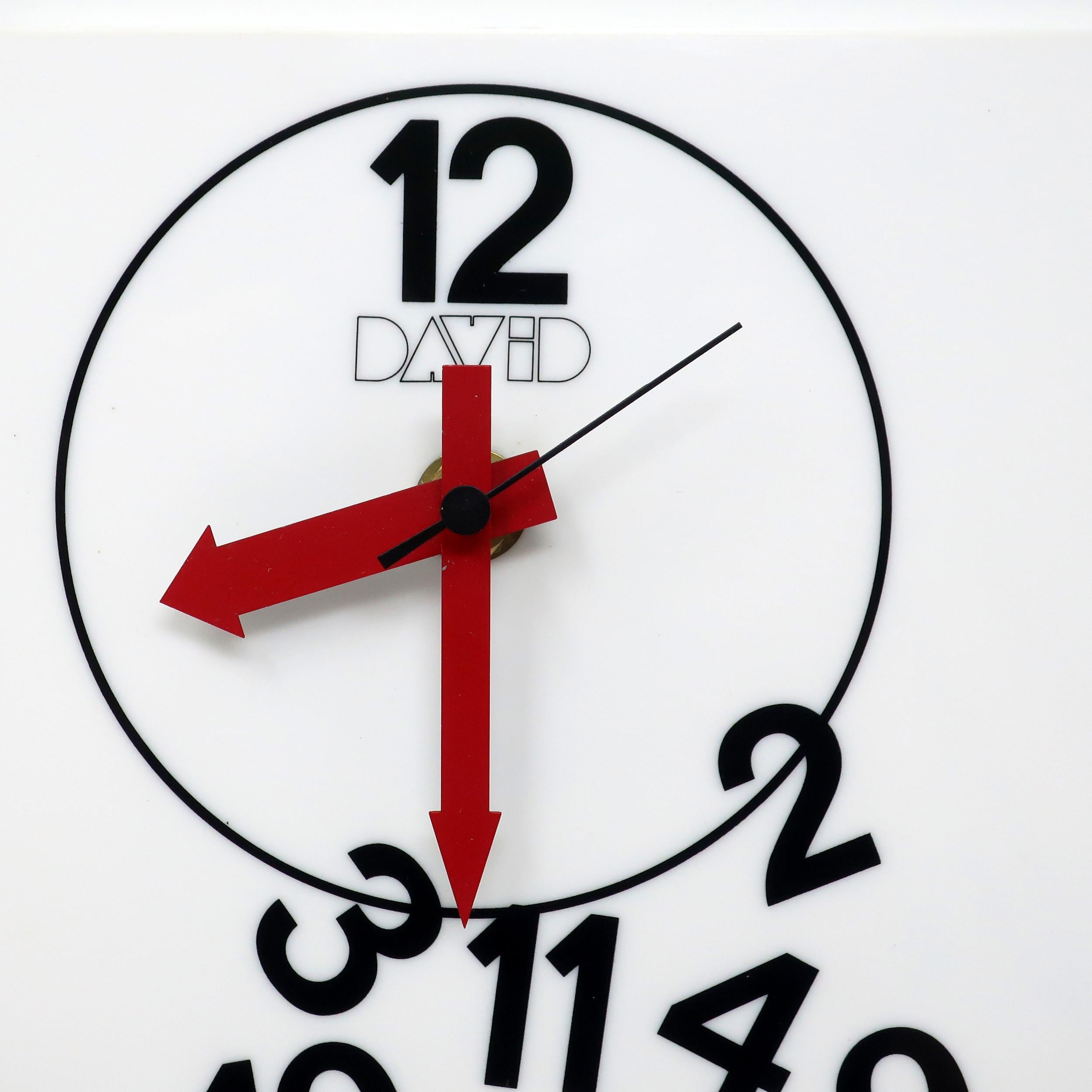 A great 1970s or 1980s wall clock designed by David Davir. A rectangular design with a white lucite face and sides, black lettering and numbers, and red arrow-shaped hands. It has all of the elements of a regular clock but the numbers (except the