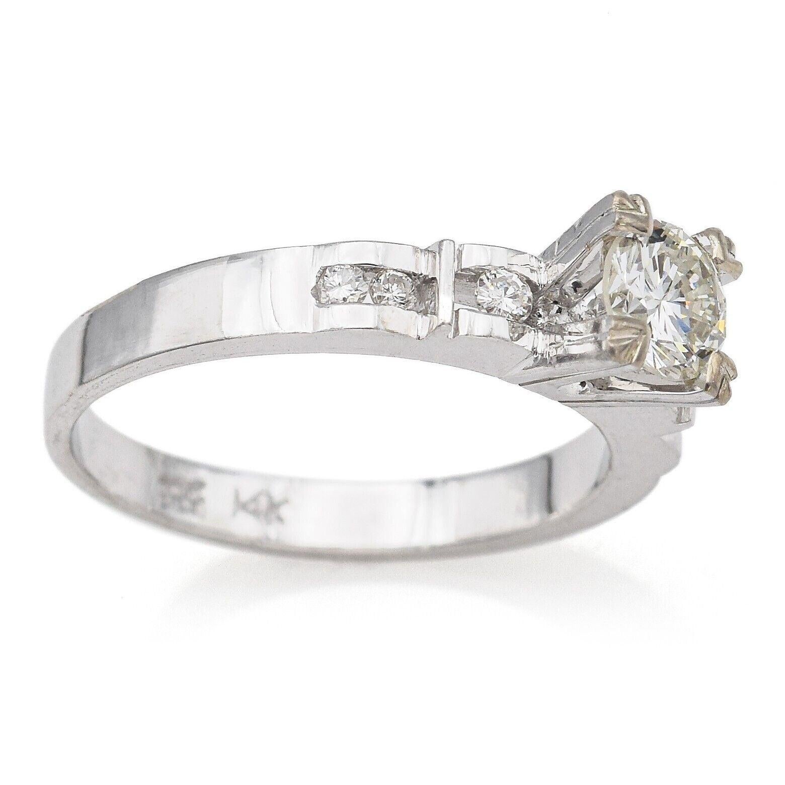 Un anello di fidanzamento vintage senza tempo realizzato in oro bianco. Al centro si trova un brillante diamante rotondo, accentuato da piccoli diamanti scintillanti incastonati lungo le spalle per una maggiore luminosità. Il design classico è