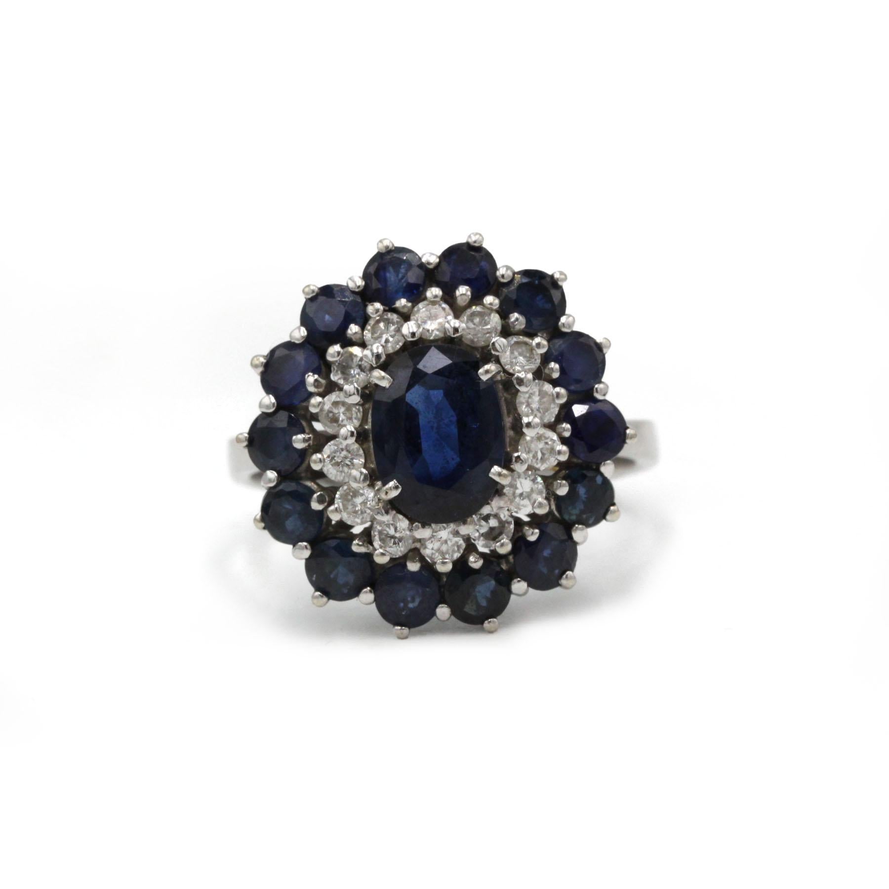 Dieser Vintage-Cluster-Ring zeigt einen tiefblauen, ovalen Saphir von 2 Karat in der Mitte, umgeben von zwei Edelsteinreihen, die eine kühne und elegante, von Blumen inspirierte Silhouette bilden. Der innere Halo besteht aus runden Diamanten im