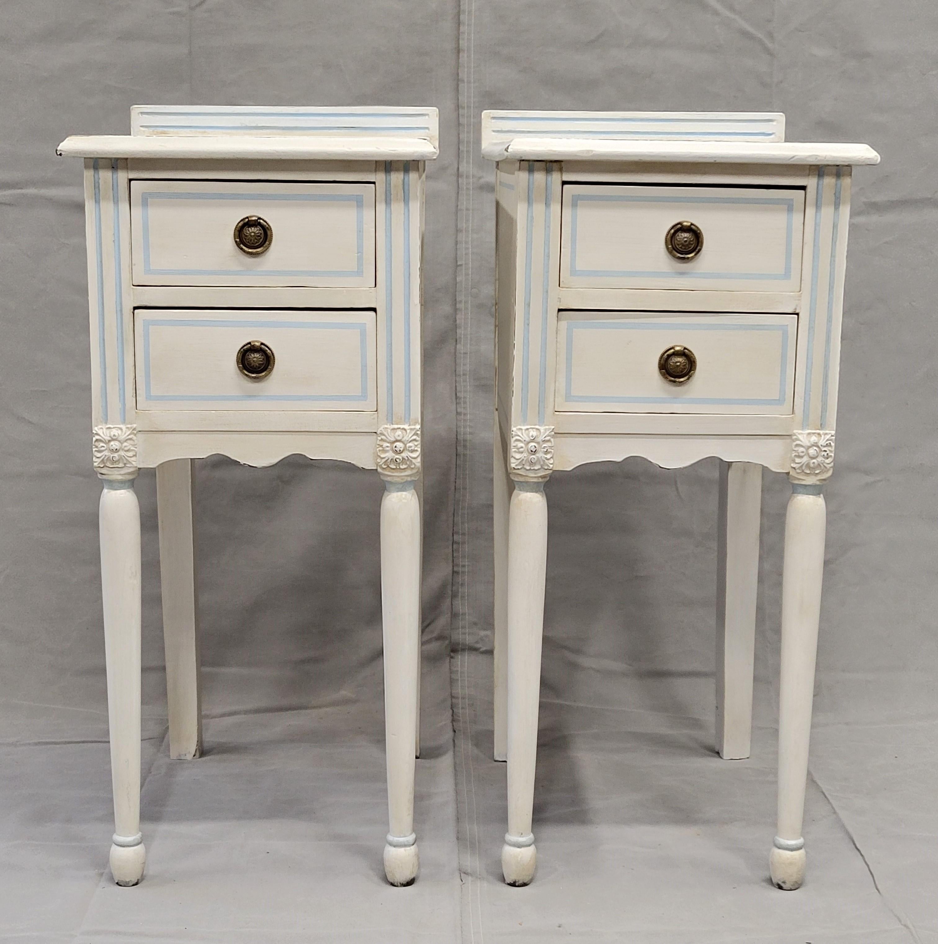 A beautiful pair of vintage nightstands painted white with pale blue pinstriping and brass pulls. Their narrow dimension makes them perfect for use in a smaller bedroom. Four drawers lined with wallpaper offer storage. The lightly distressed painted
