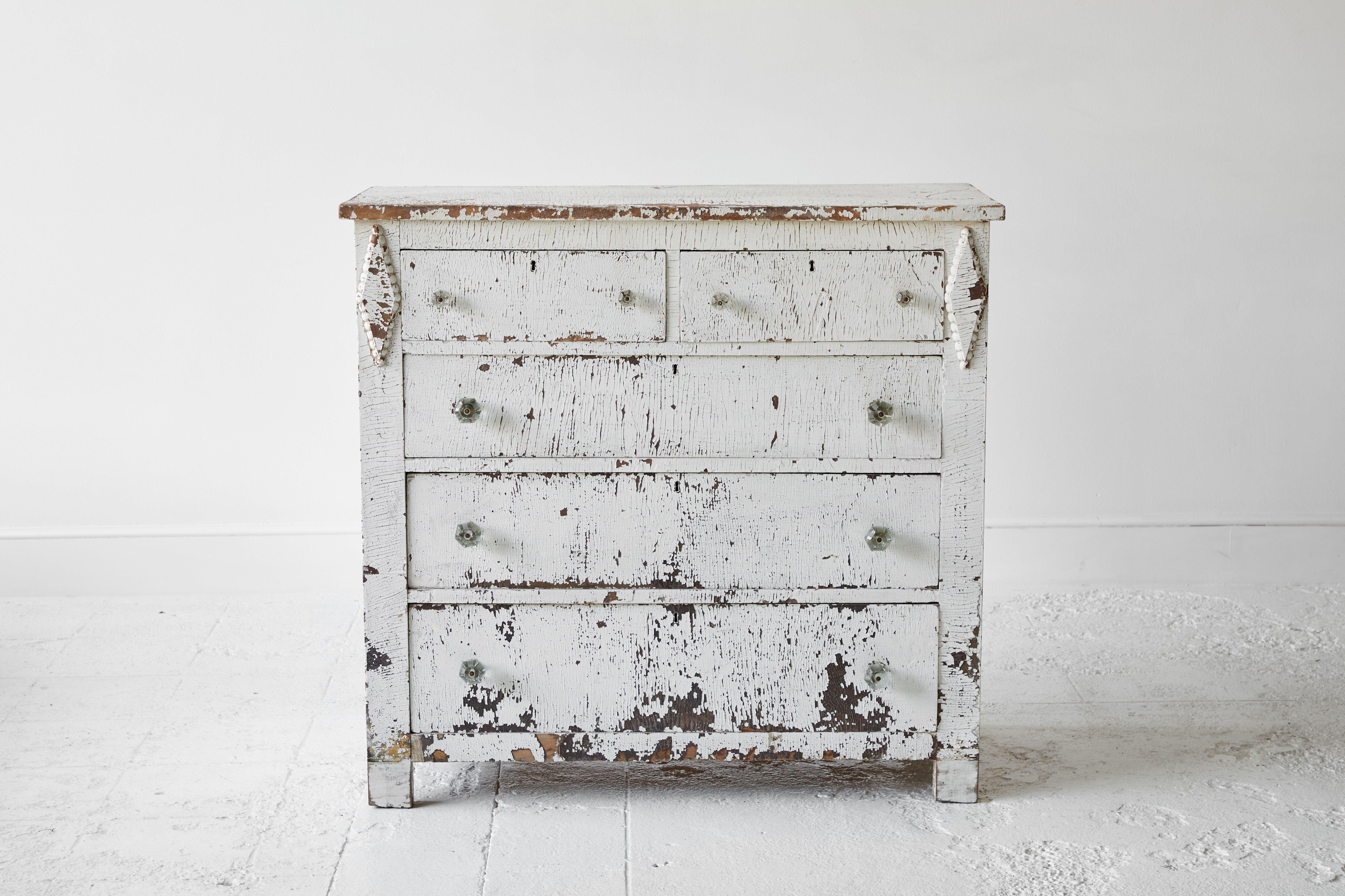 Five-drawer white painted chest of drawers flanked with two diamond motifs. The paint is chipping which offers lots of character and patina.
