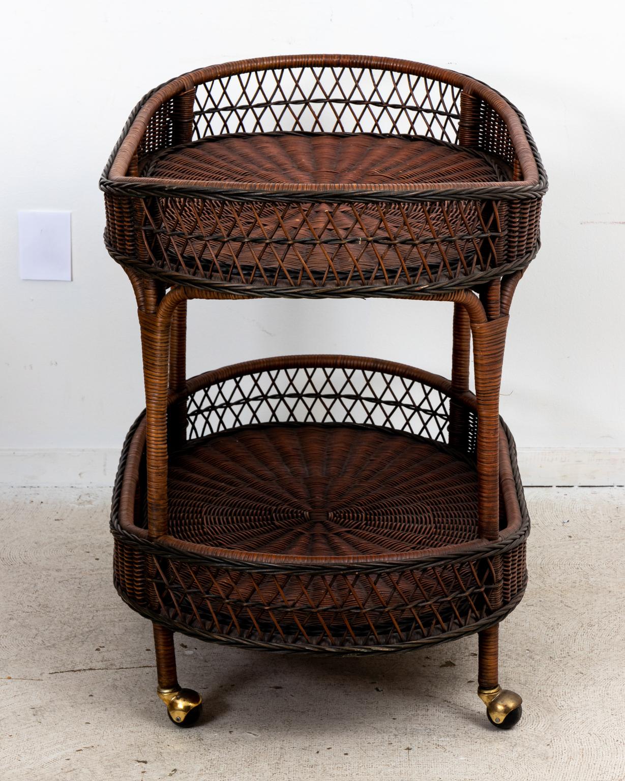 Circa 1940s two tier bar cart on brass wheels with braided wicker rattan trim. Please note of wear consistent with age including minor finish loss to the wicker and patina to the brass.