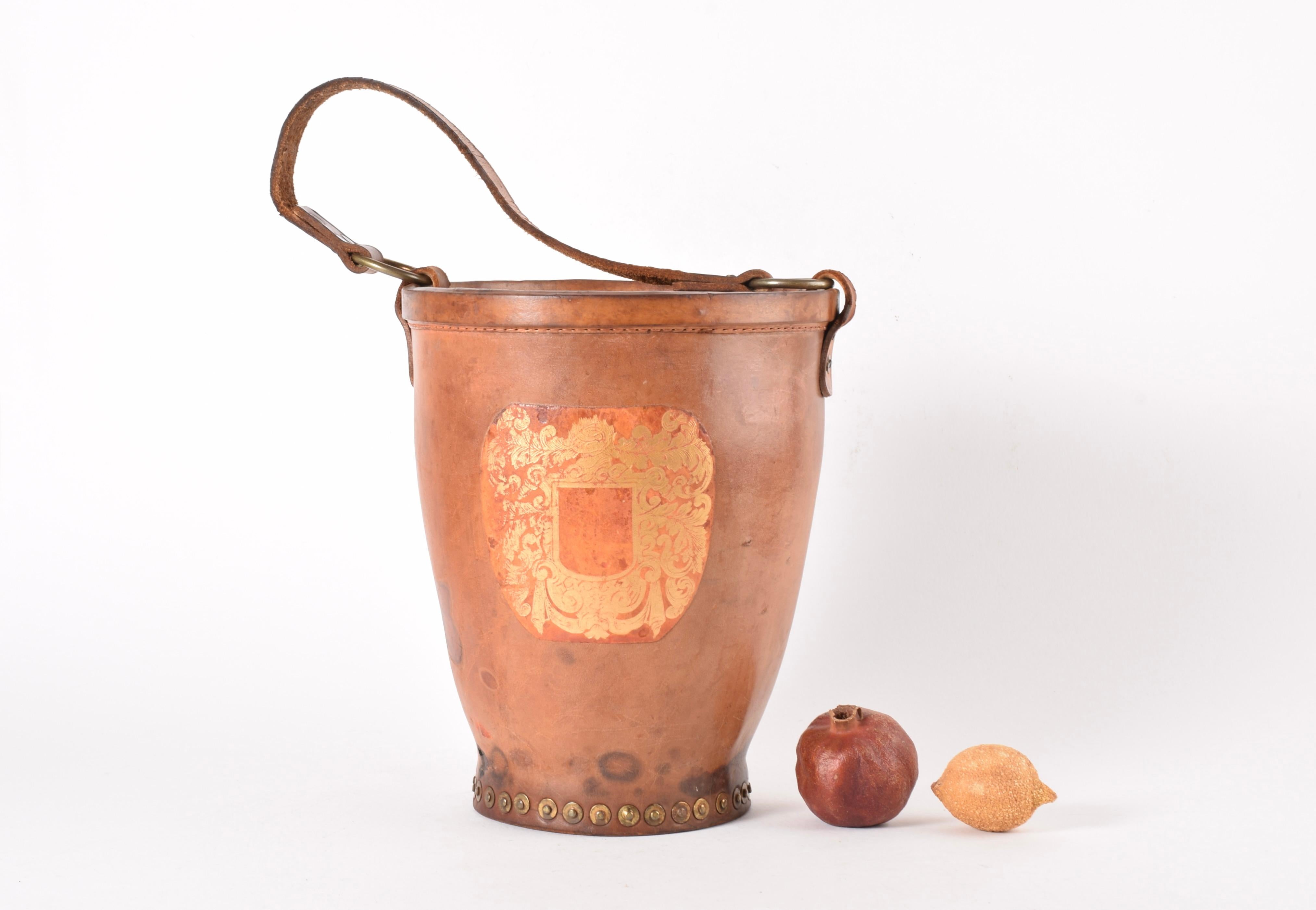 Cubitera o enfriador de champán/vino vintage fabricado en cuero de plena flor. Fabricado en Inglaterra. Suponemos que es de hacia 1920.

Está montado con muchos remaches decorativos de latón y tiene una decoración floral impresa con oro sobre cuero